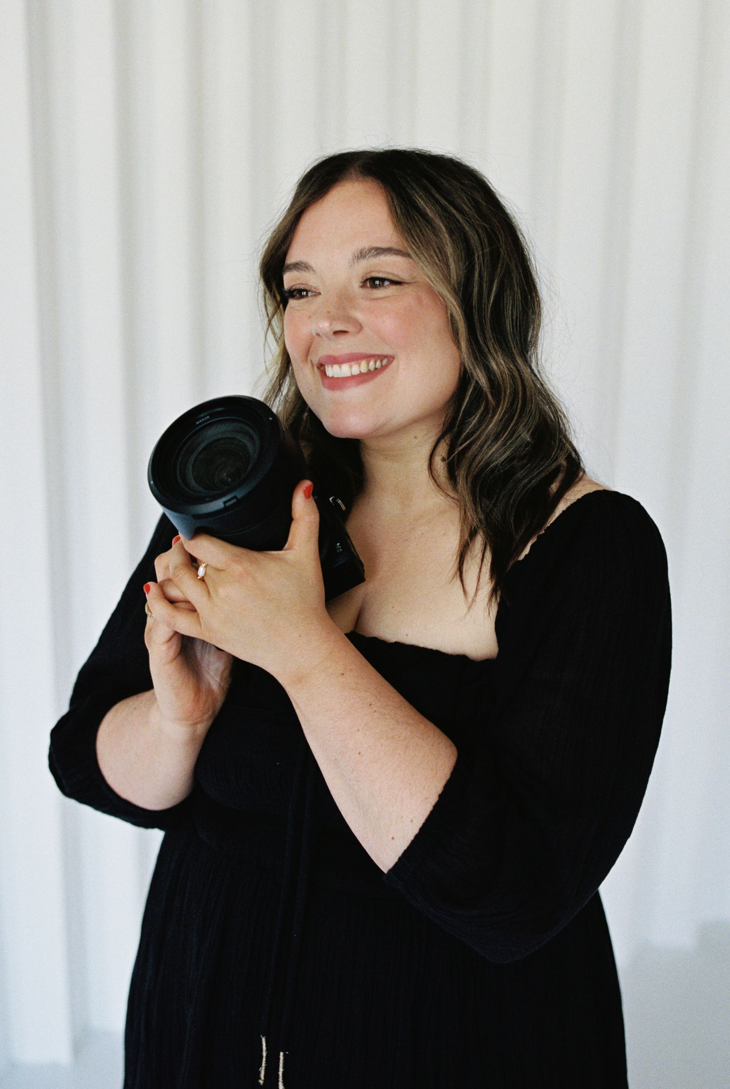Smiling woman in black dress holding a professional camera, posing against a white background.