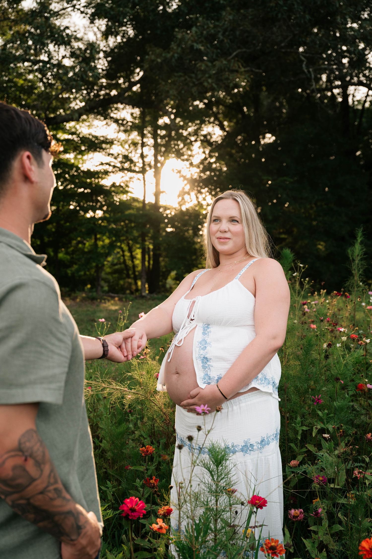 Pregnant woman in white dress holds hands with tattooed partner in a wildflower field at golden hour sunset.