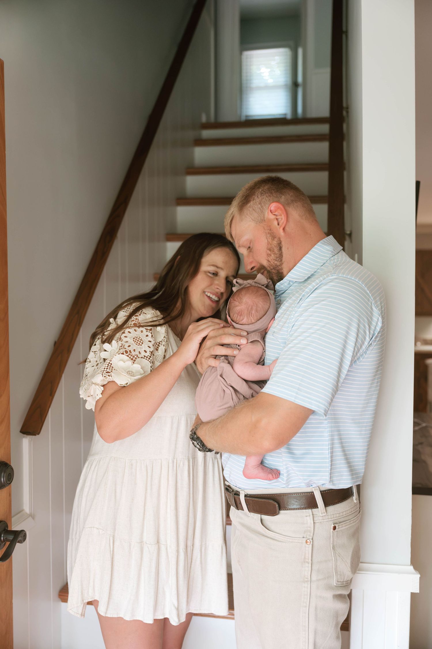 A couple shares a tender moment with their newborn baby while standing by a staircase in their home.