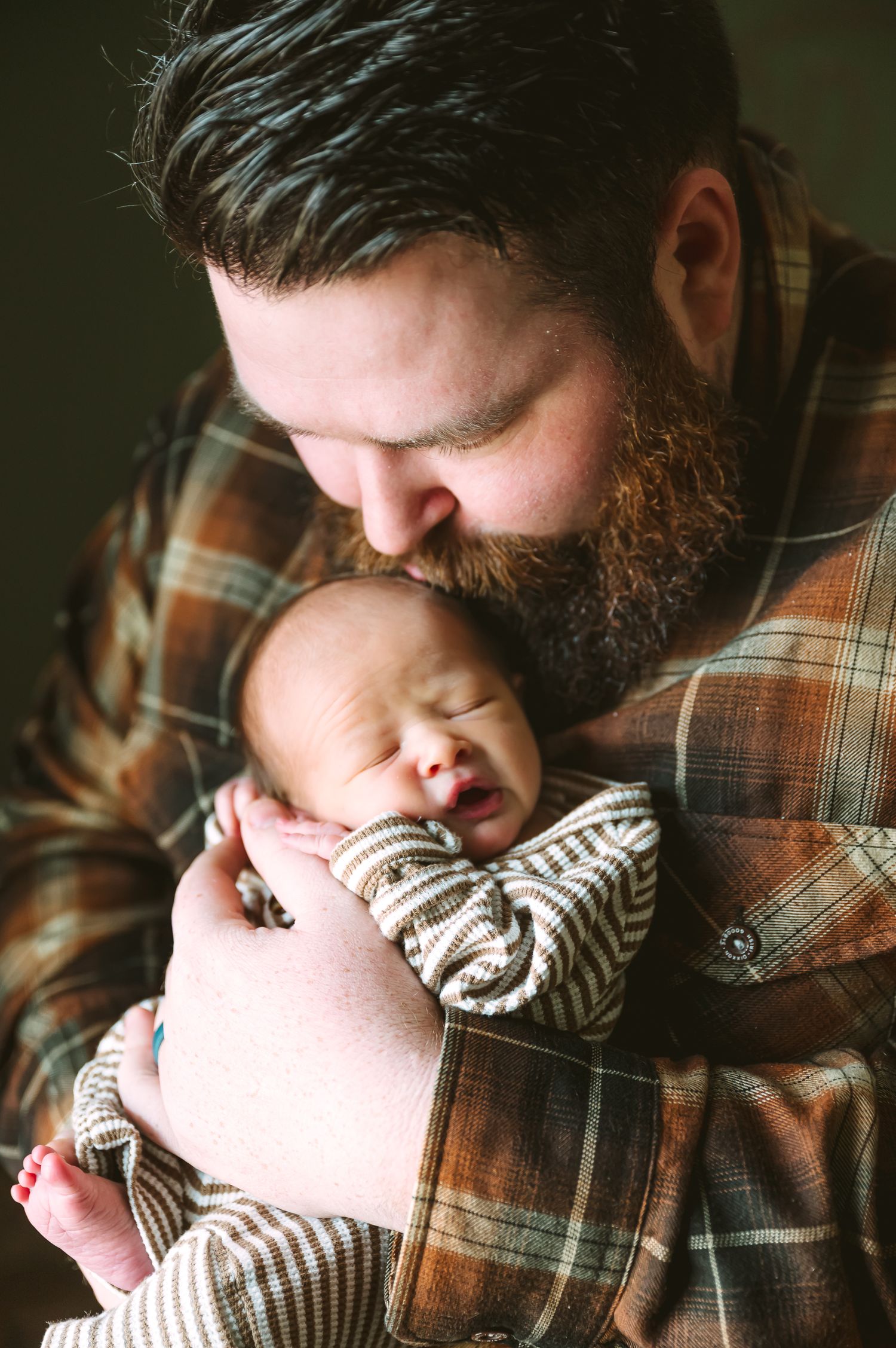 Intimate portrait of parent and baby in plaid shirt against dark background.