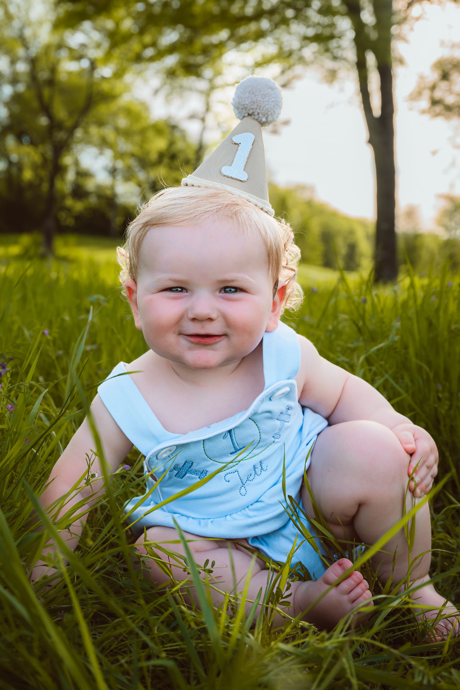 A baby wearing a blue outfit and party hat sits smiling in tall grass.