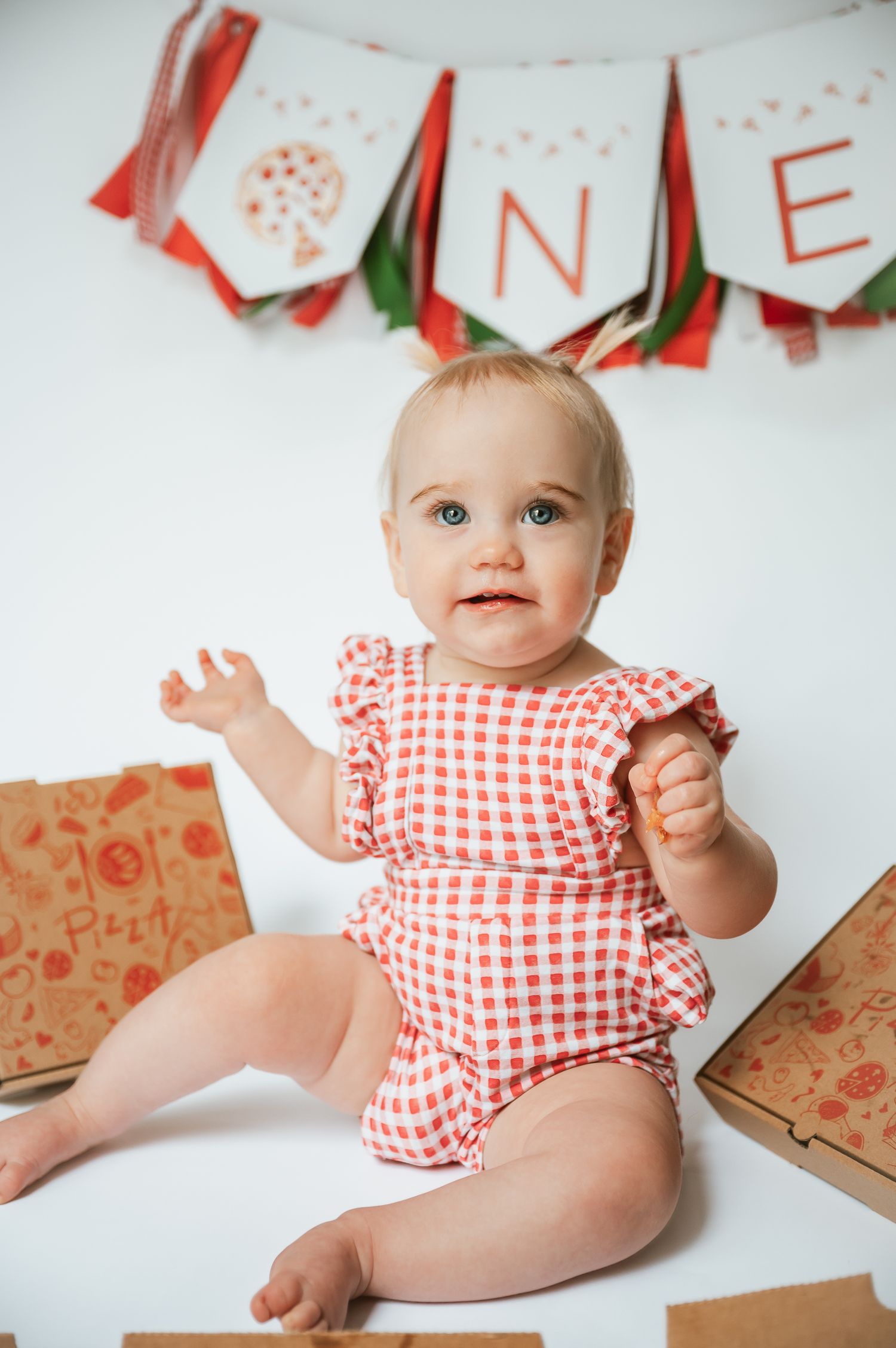Baby in red gingham romper sitting under a festive banner.