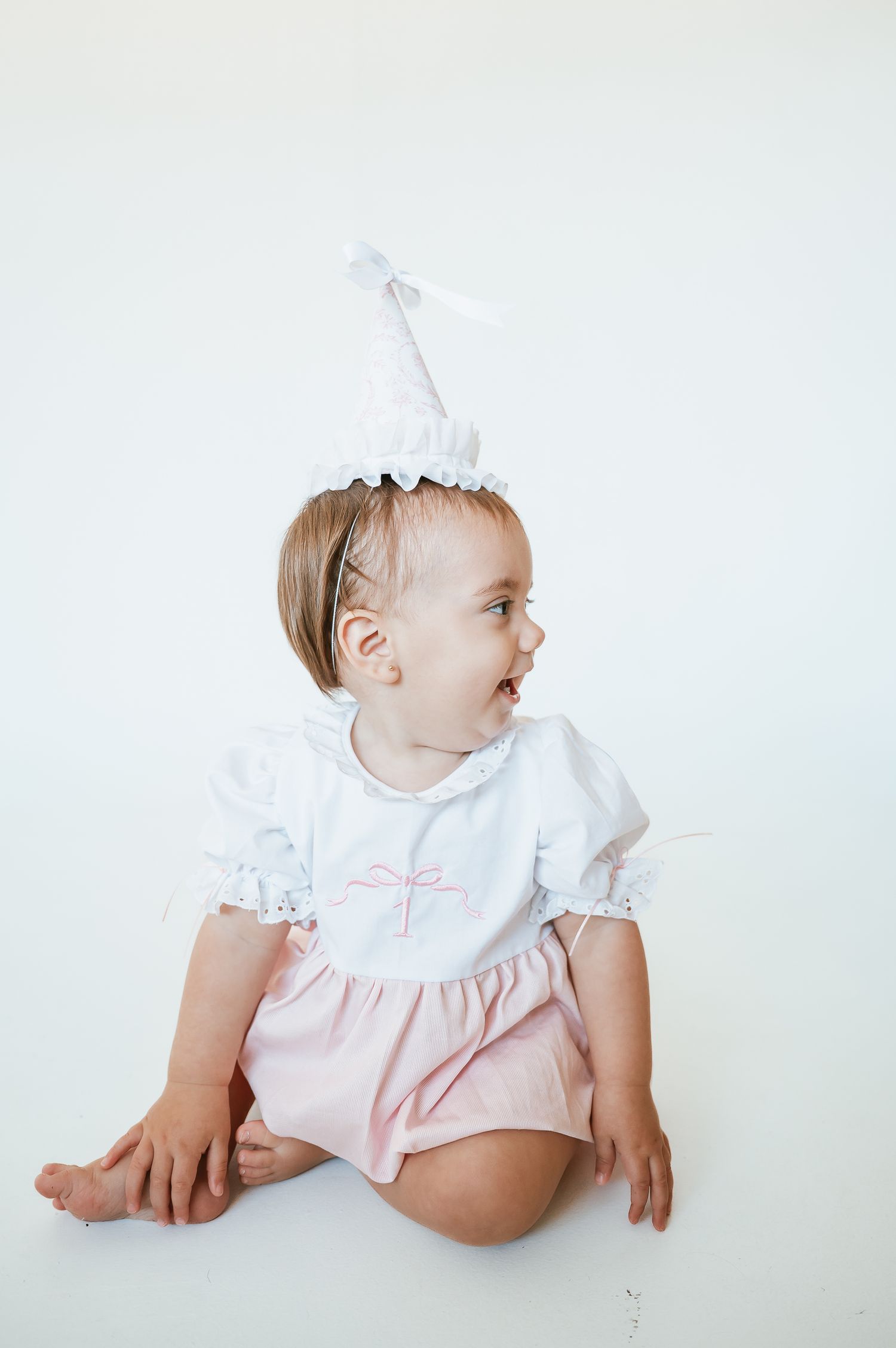 Baby in white dress photographed against white background.