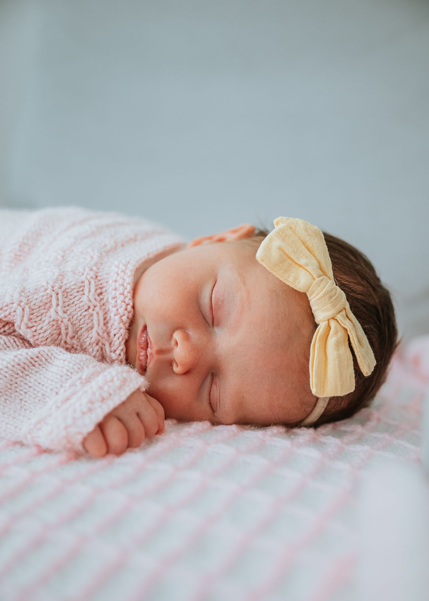 A sleeping newborn with yellow bow headband wrapped in pink lace blanket.