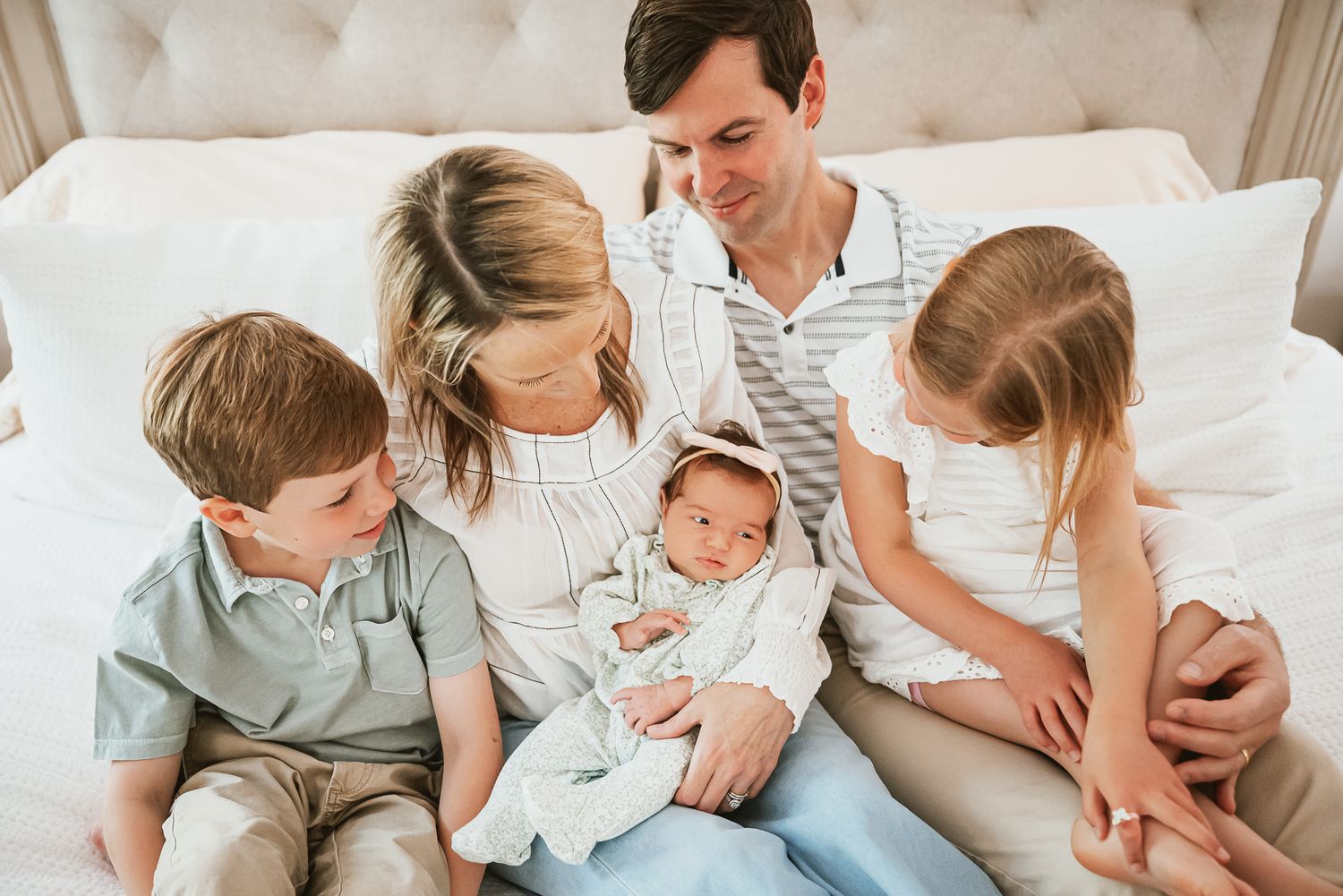 A family of five sits together on a beige tufted bed with their newborn baby.