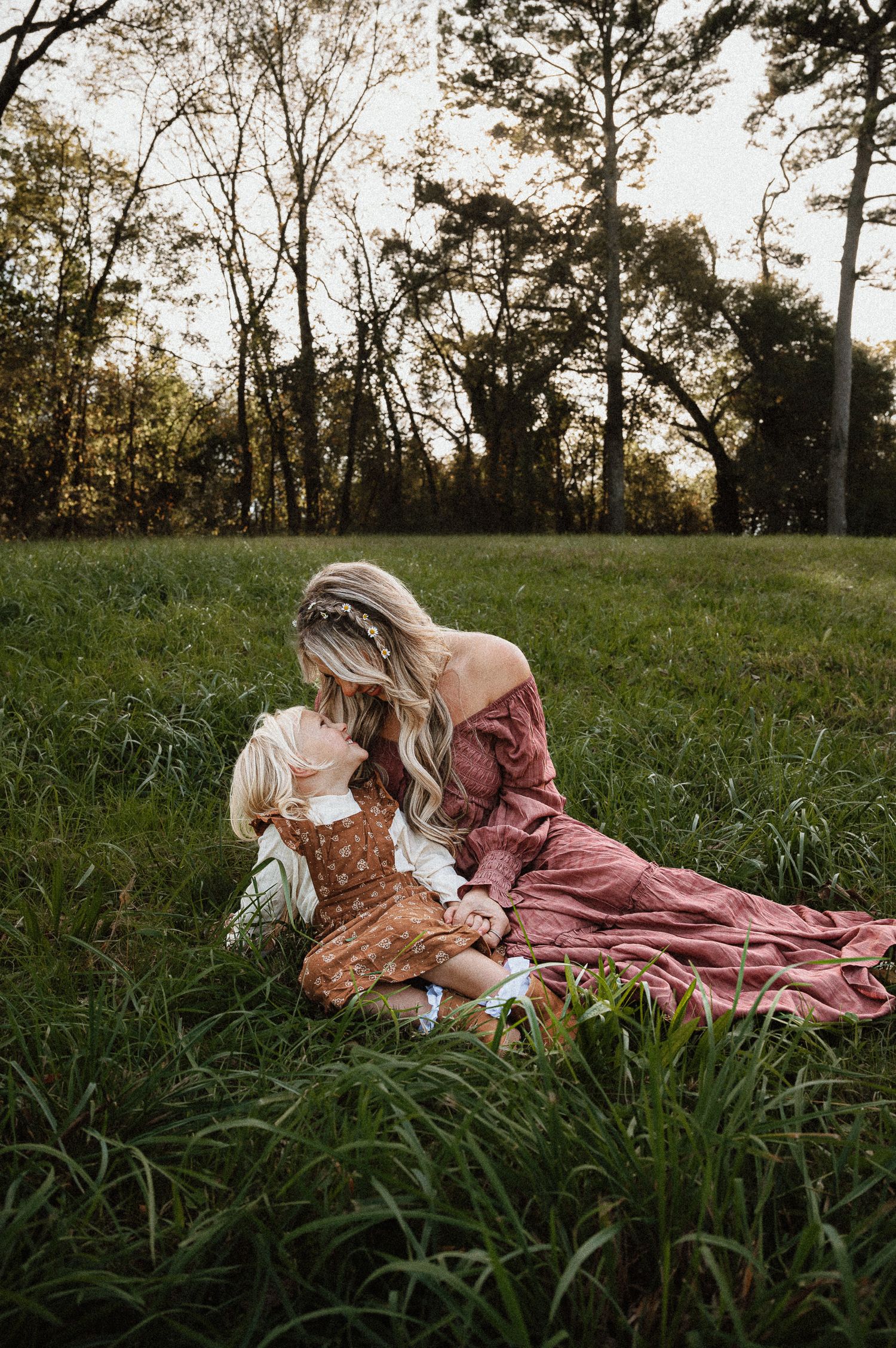 A mother and daughter in vintage dresses sit together in a sunlit meadow at golden hour.