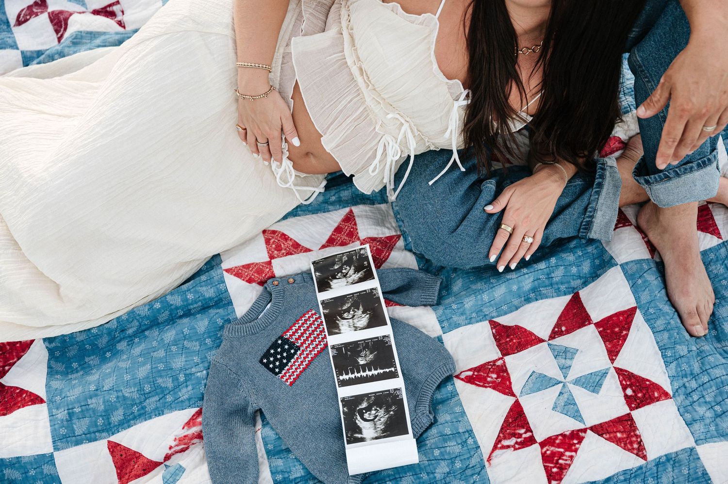 Two women sit on a patriotic quilt with baby ultrasound photos, tiny denim outfit, and American flag for pregnancy announcement.