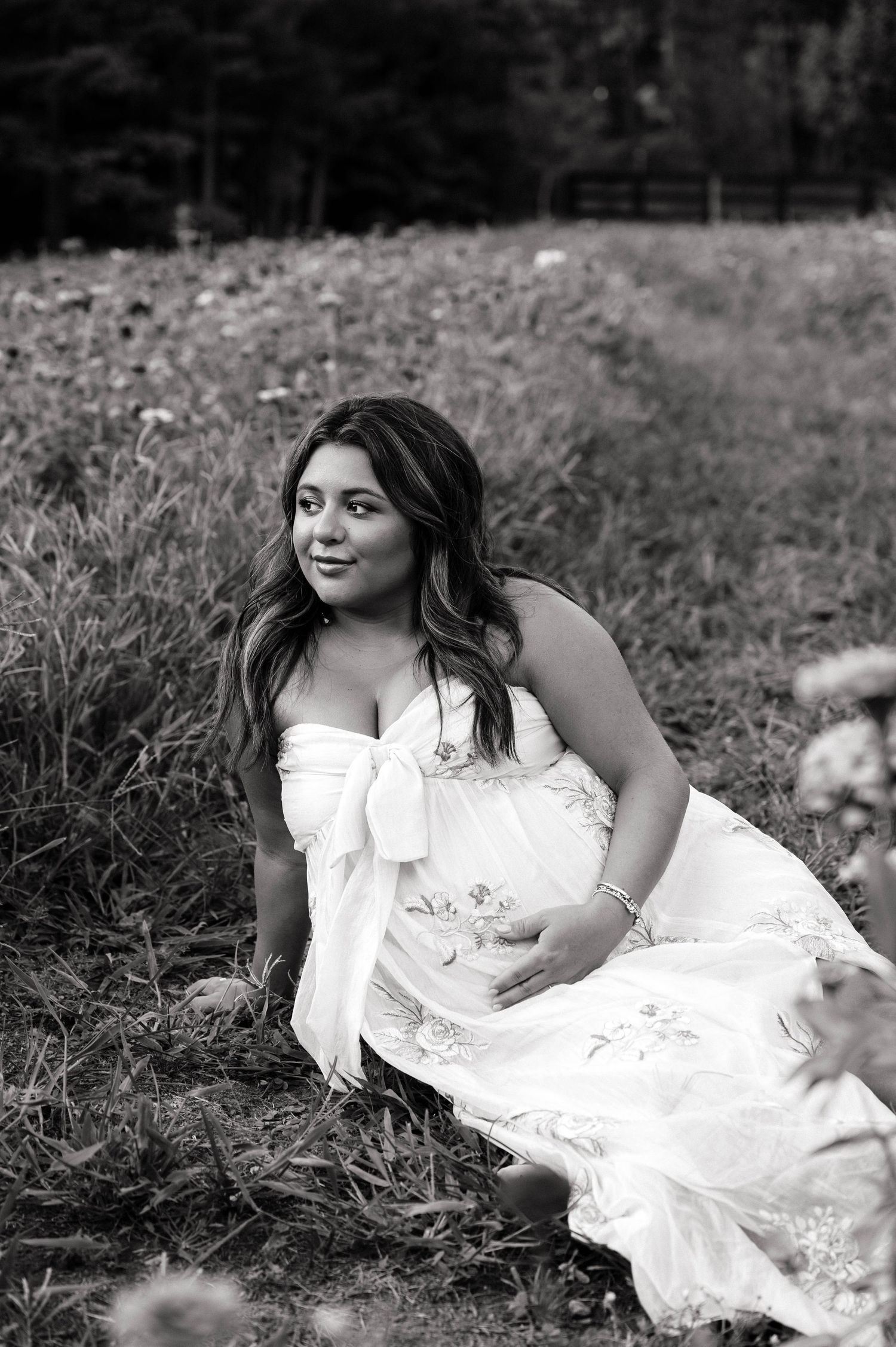 Black and white photo of a pregnant woman in a white floral dress sitting in a grassy field, smiling softly.