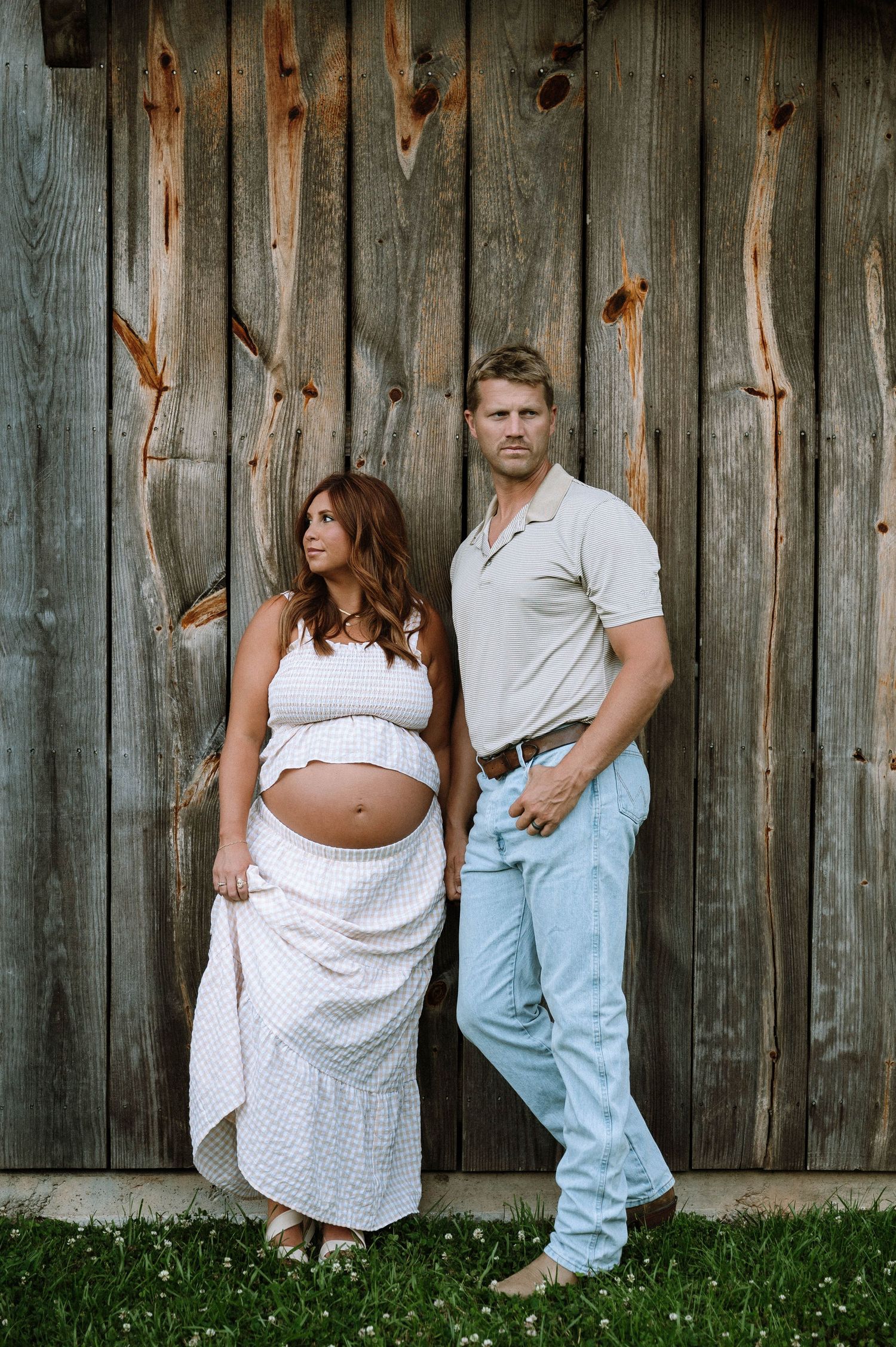Pregnant couple posing against rustic wooden barn wall, woman in white crop top and skirt showing baby bump, man in light jeans.