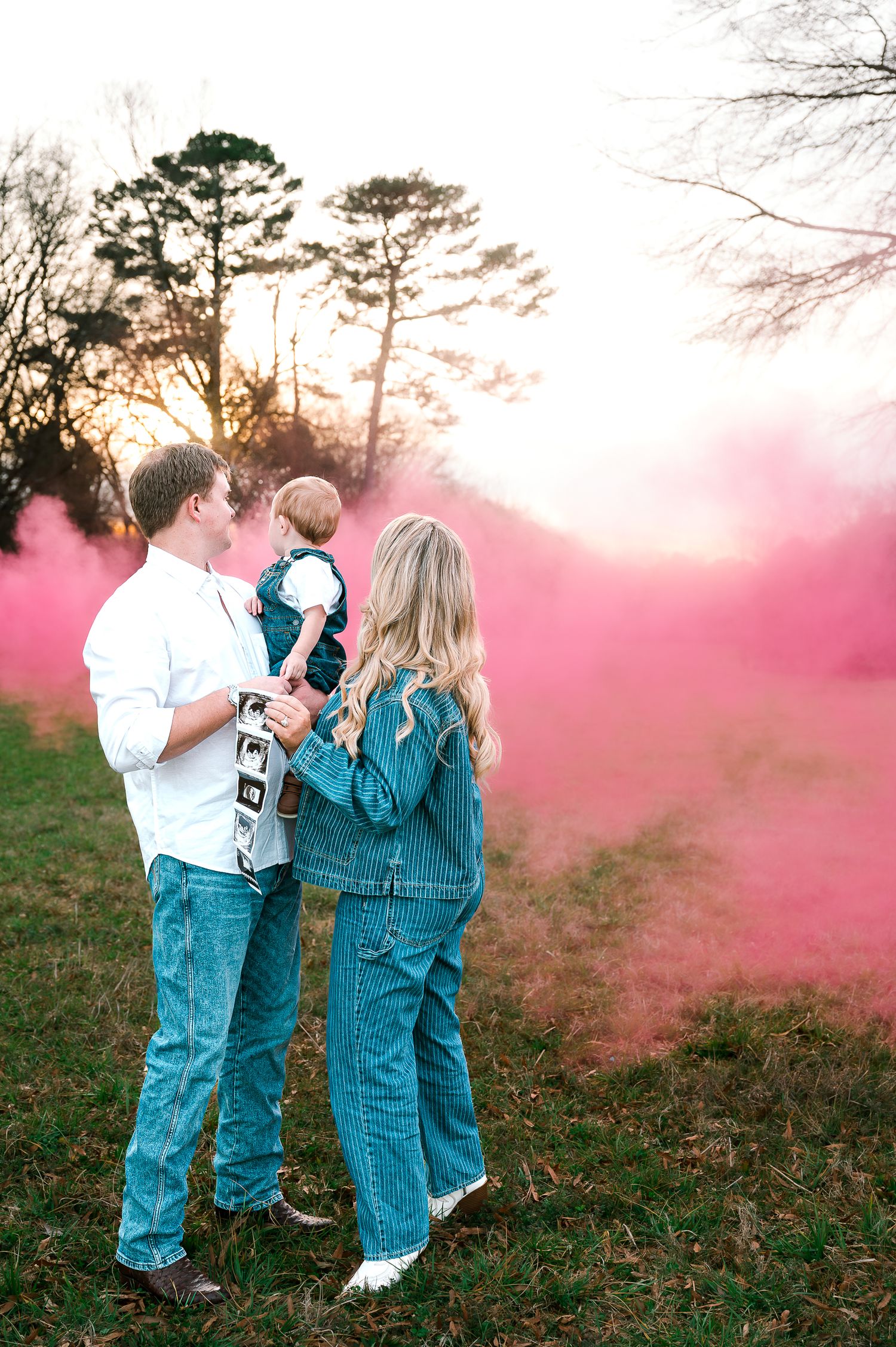 Family gender reveal with pink smoke bomb at sunset, parents in denim holding baby in grassy field.