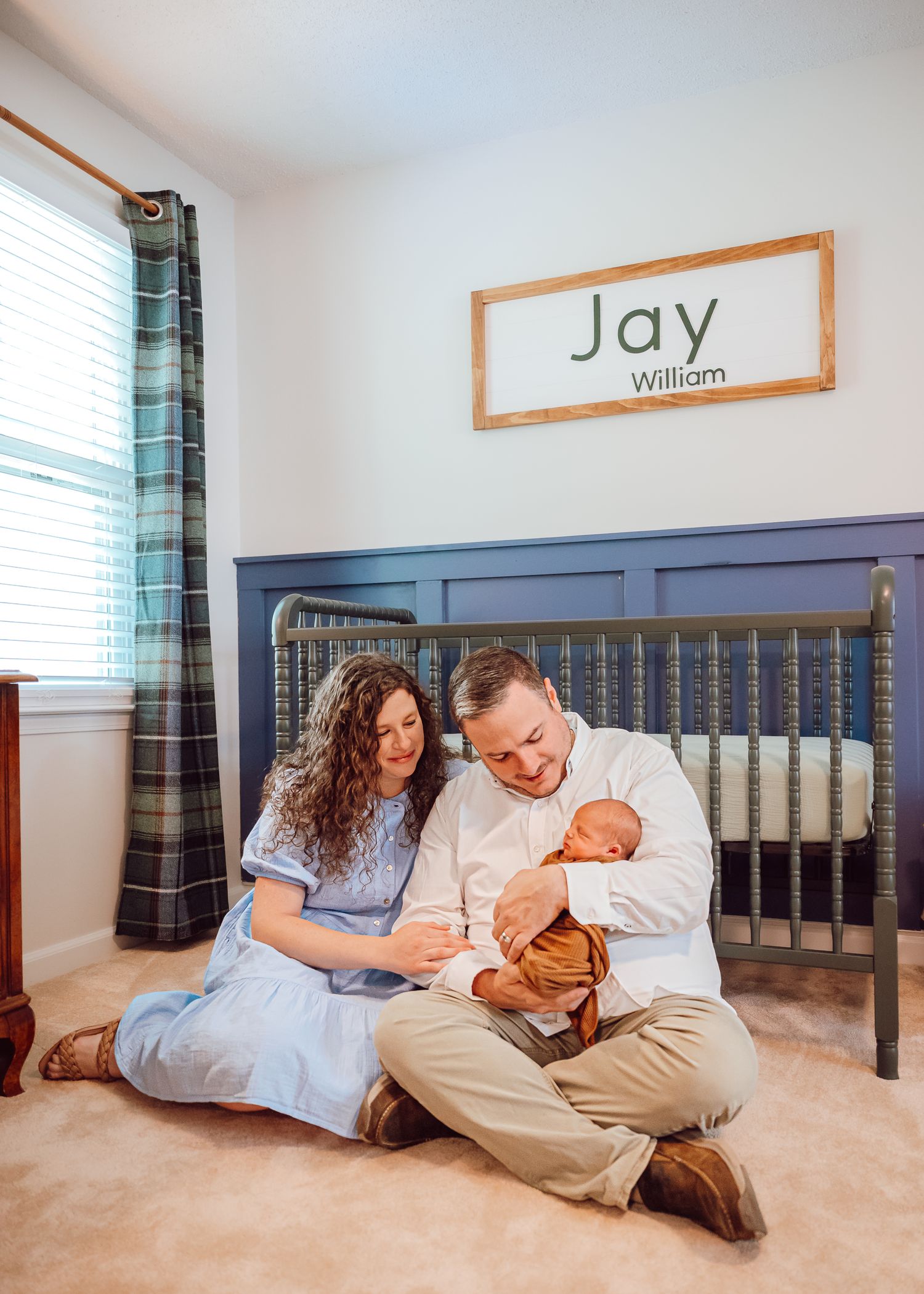 Parents sit on nursery floor with newborn under wooden name sign reading Jay William.