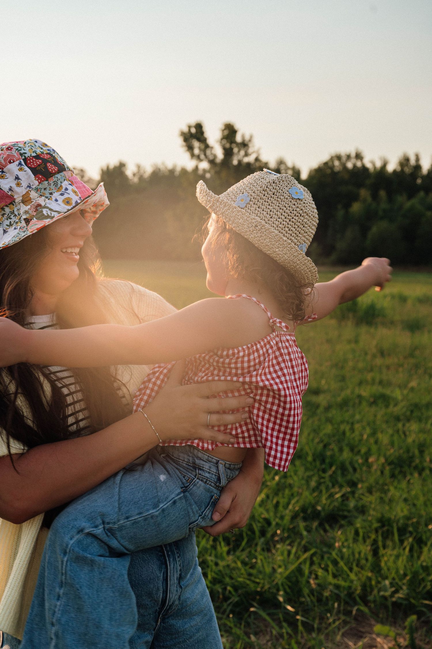 A smiling woman in a floral hat holds a young girl in a straw hat in a sunny green field.