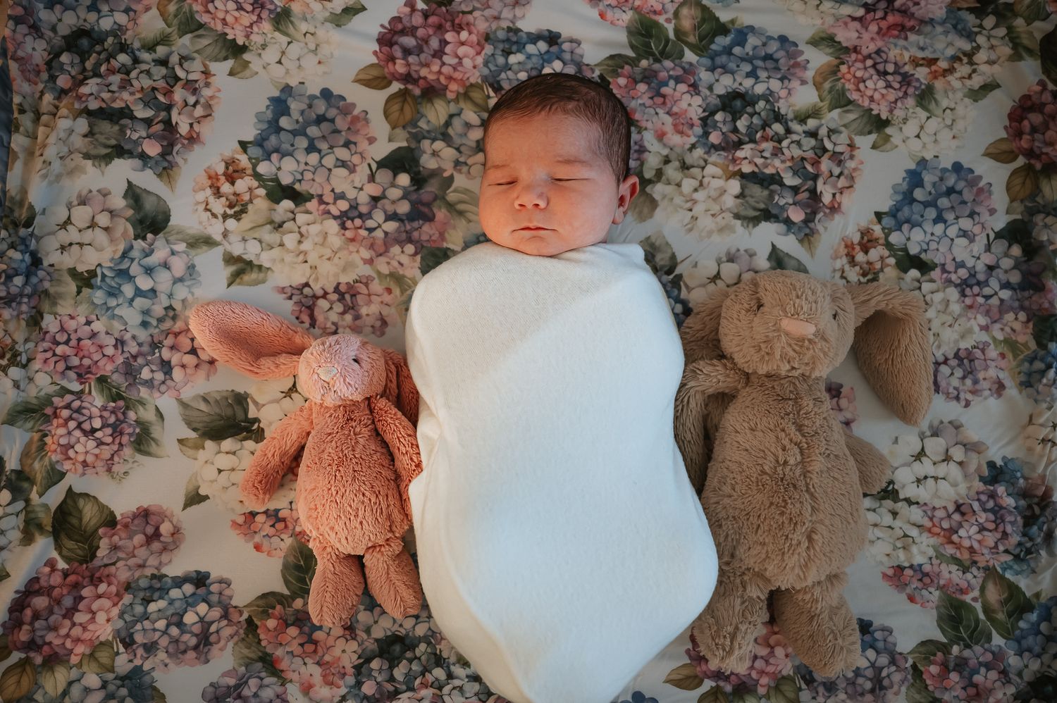 Sleeping newborn swaddled in white between two plush teddy bears on a floral blanket.