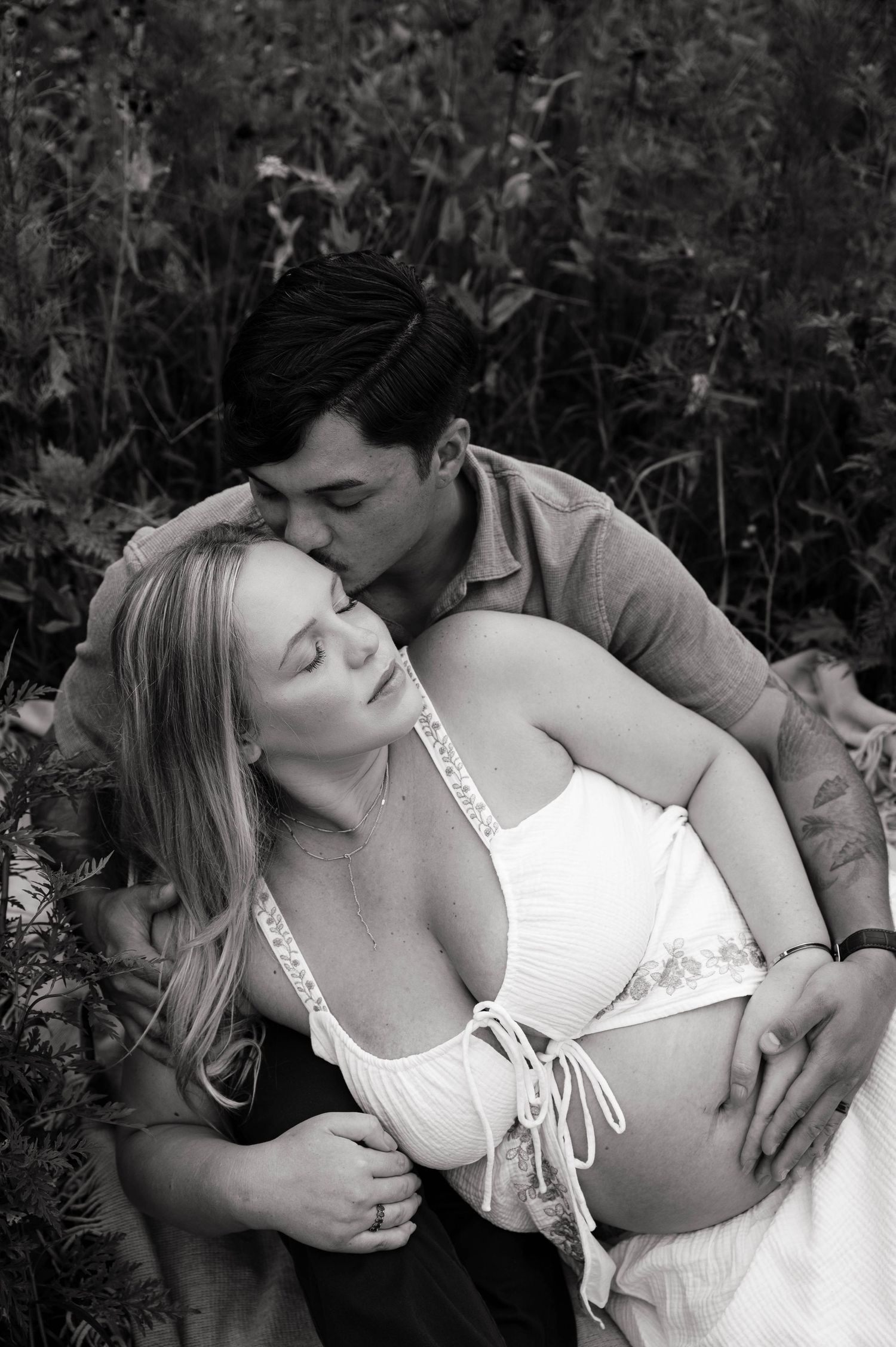 Black and white maternity photo of a couple outdoors, man kissing woman's head while cradling her pregnant belly.