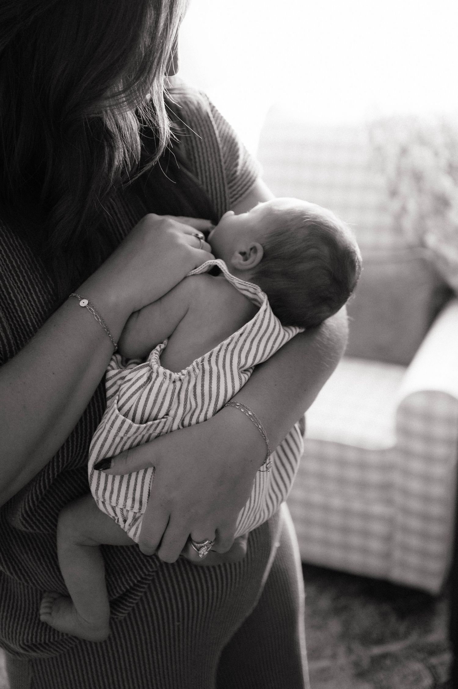 Black and white photo of a mother cradling her newborn baby, wearing a bracelet and ring, with a cozy chair in the background.