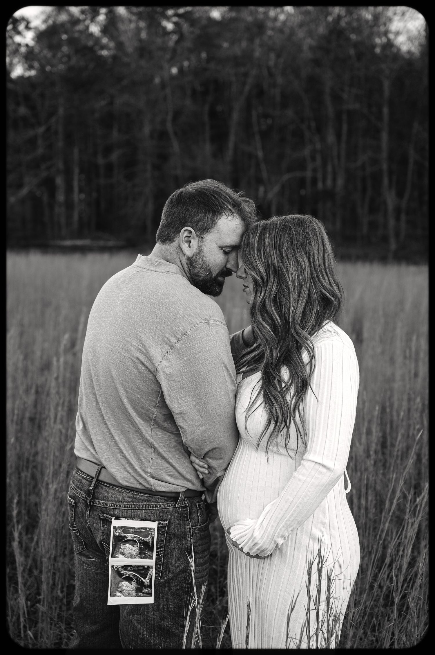 A couple embraces in a field while holding ultrasound photos in black and white.