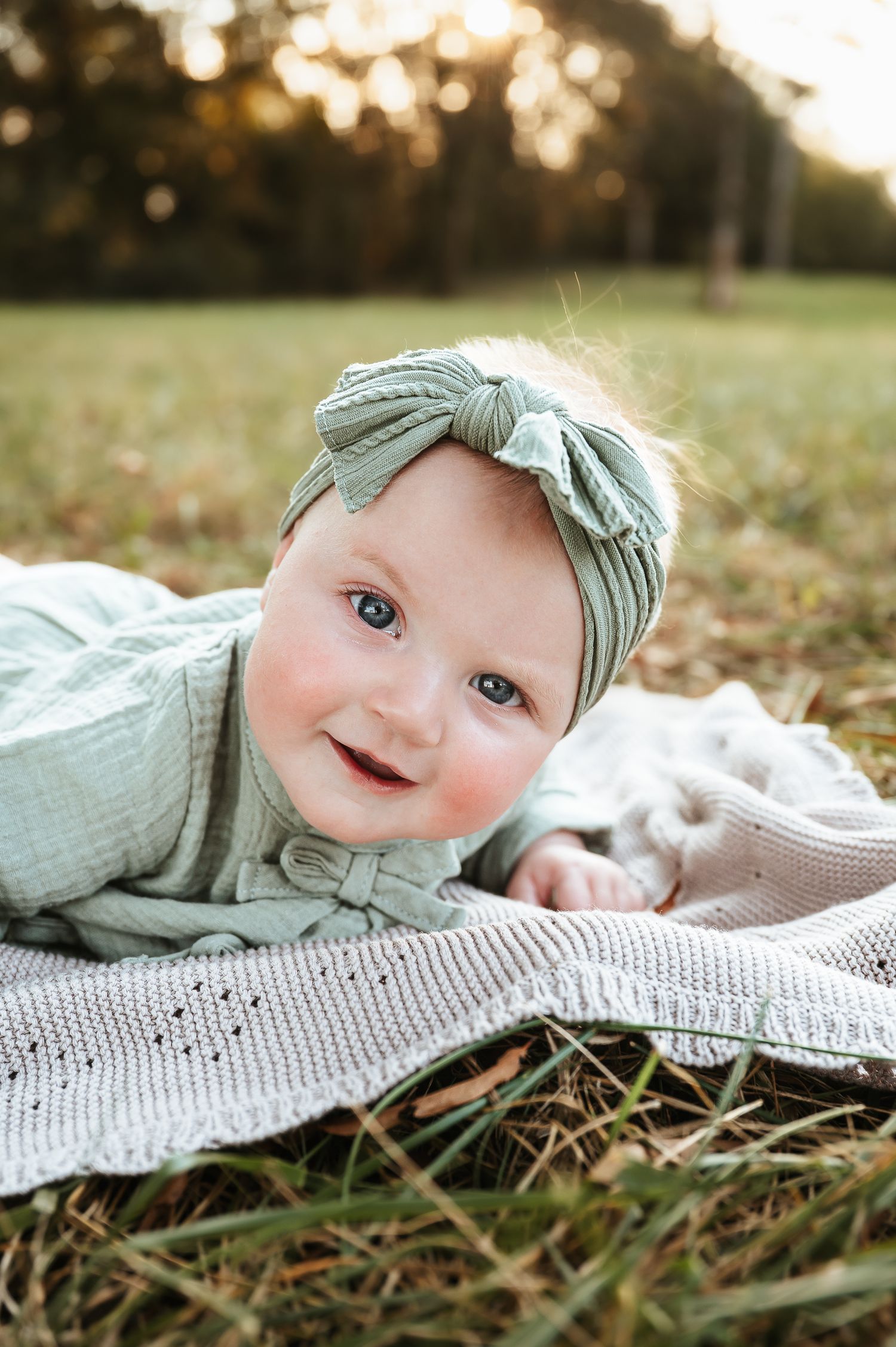 Baby in mint green outfit lying on blanket outdoors during golden hour.