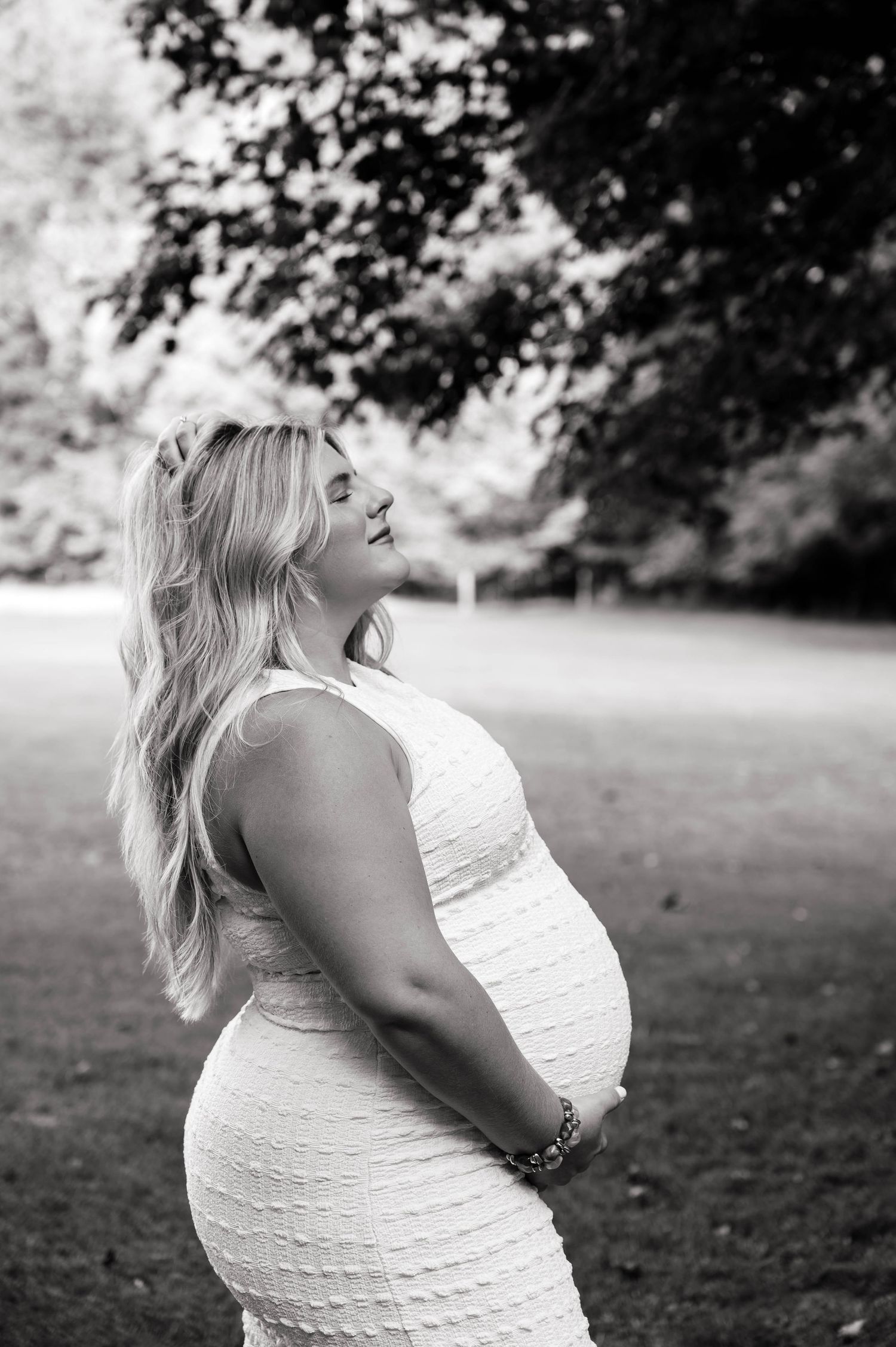 Black and white photo of a pregnant woman in a white dress holding her baby bump outdoors in a park.