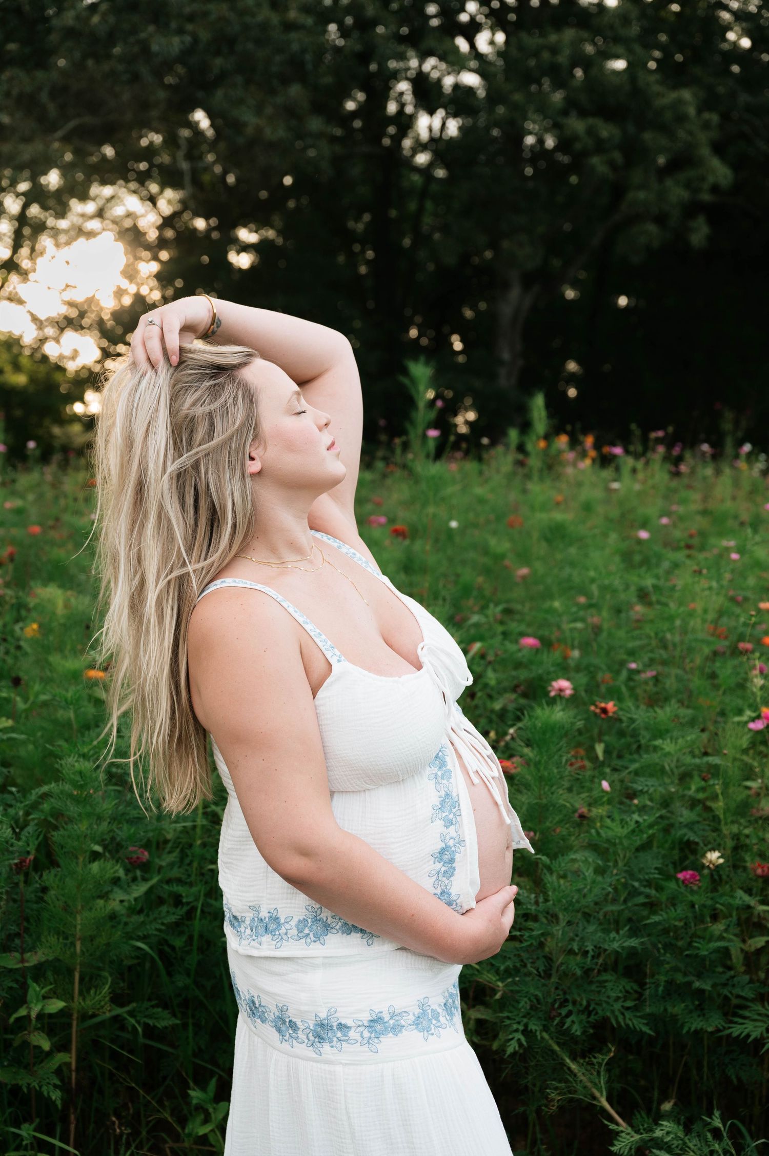 Pregnant woman in white floral dress holding belly in a blooming wildflower field at golden hour.