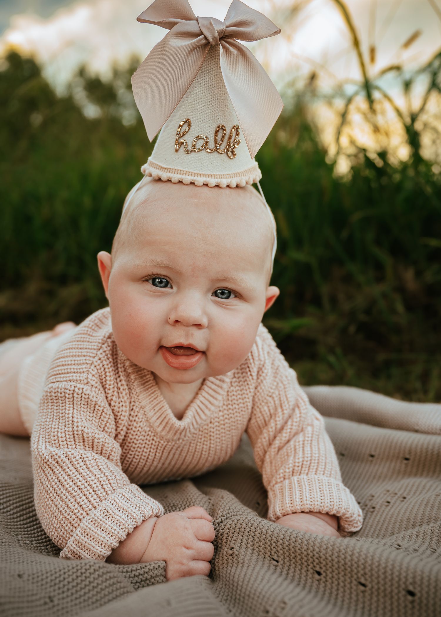 Baby in knit sweater with bow lying on textured blanket during sunset.