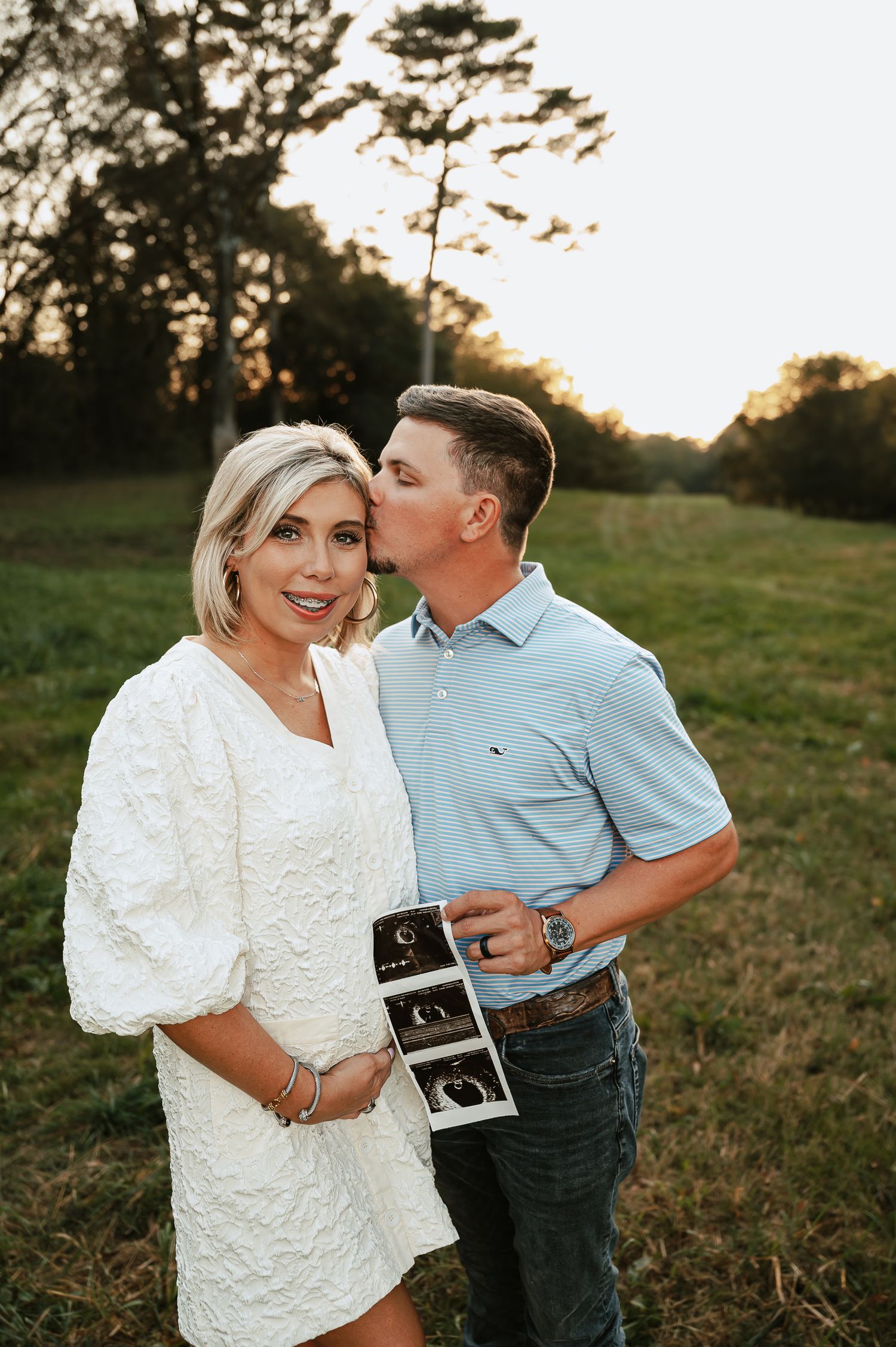 A couple shares a tender moment outdoors while holding ultrasound photos during golden hour.