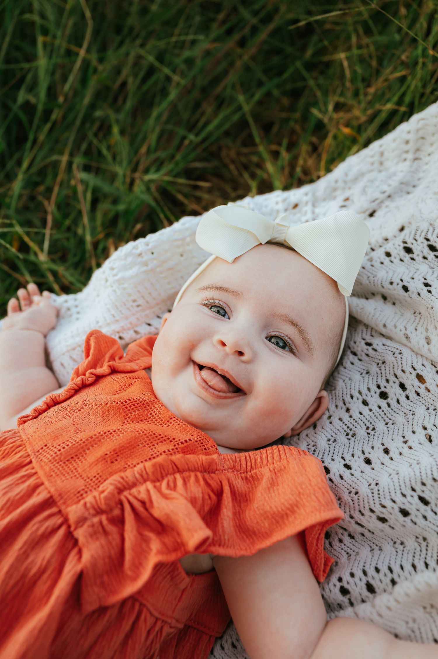 Baby in orange ruffled dress lying on white lace blanket in grass.