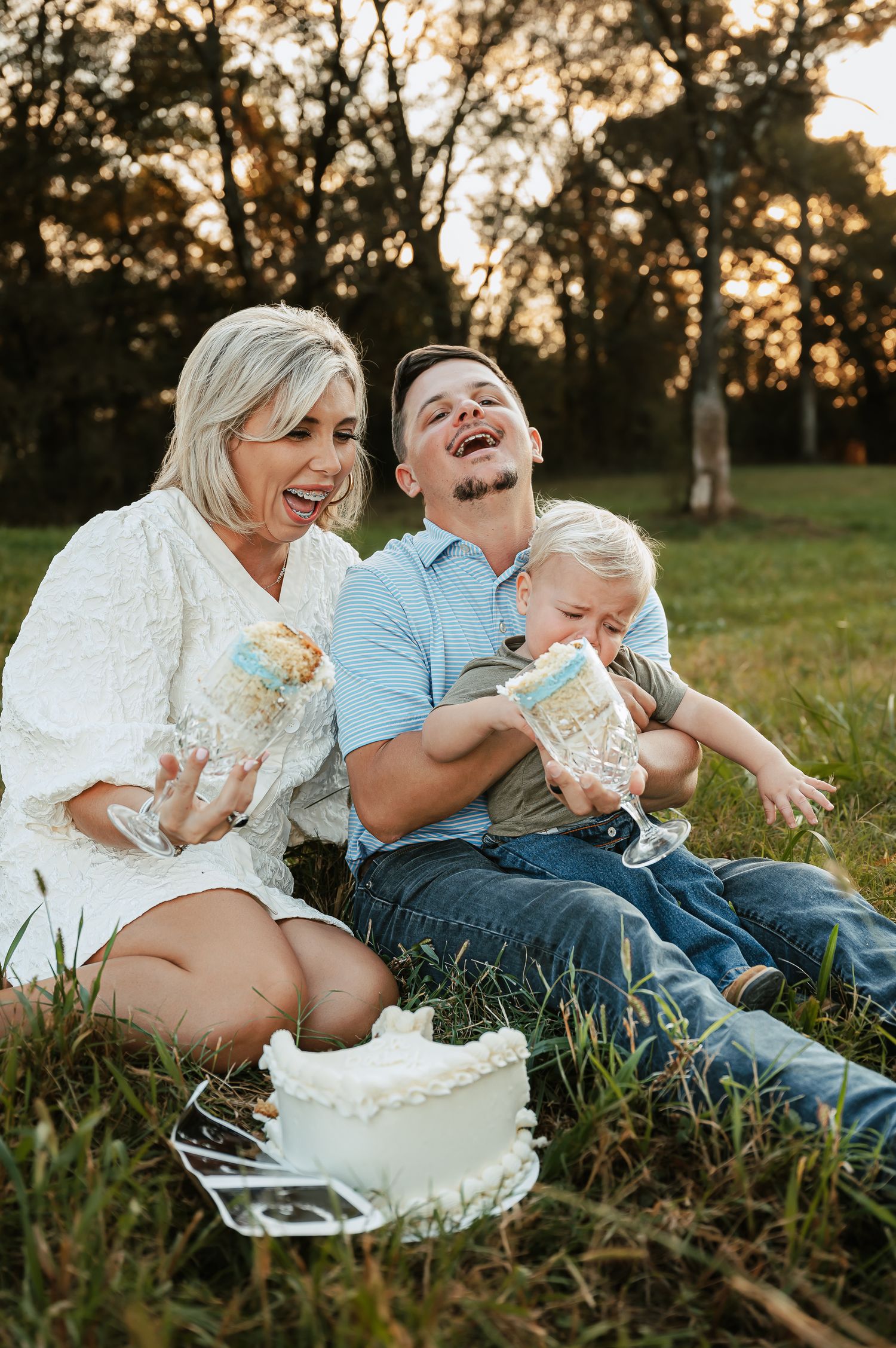 A family laughs together while having cake during an outdoor photoshoot at sunset.