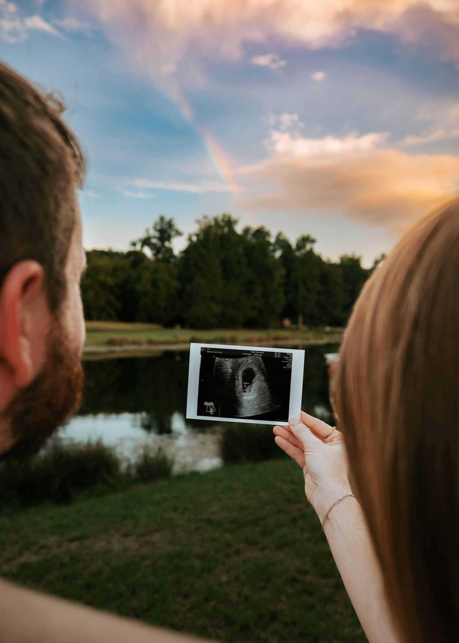 Couple holding ultrasound photos against sunset sky by a lake.