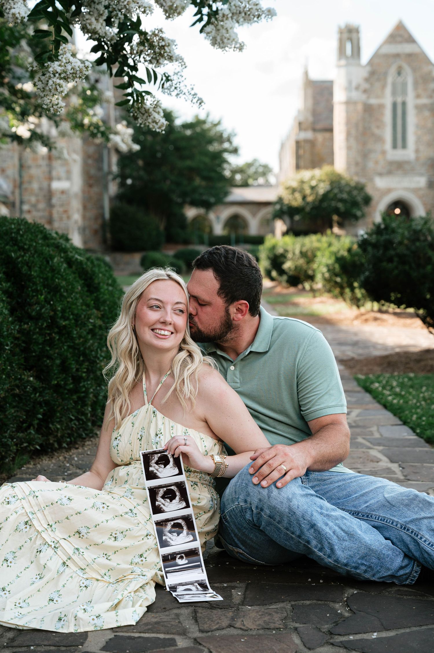 Happy couple sitting outdoors holding ultrasound photos, sharing a kiss during a pregnancy announcement photoshoot.