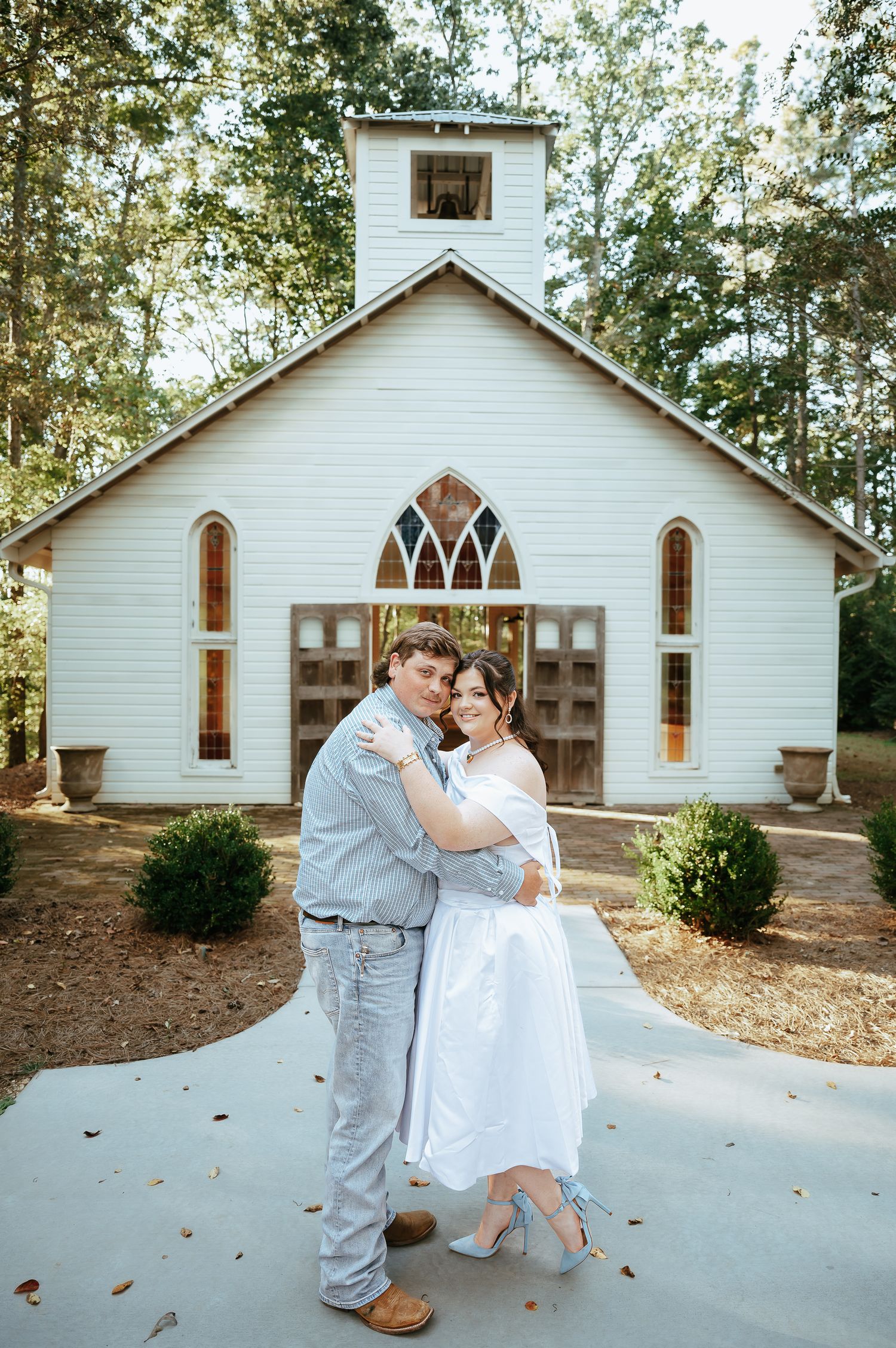 Romantic couple embracing outside white chapel with gothic windows and wooden doors.