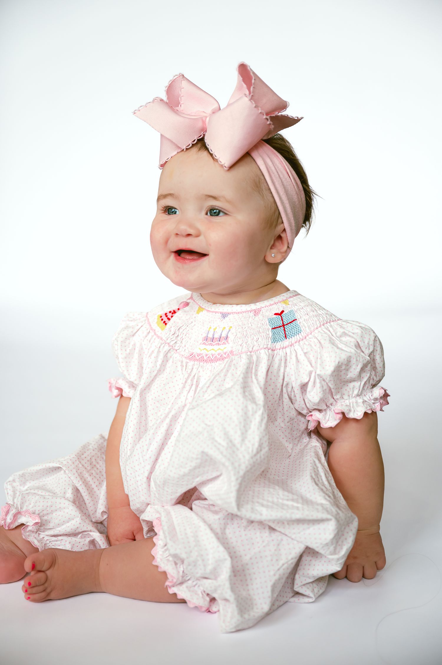 Baby in white smocked romper and pink bow headband smiling against white background.