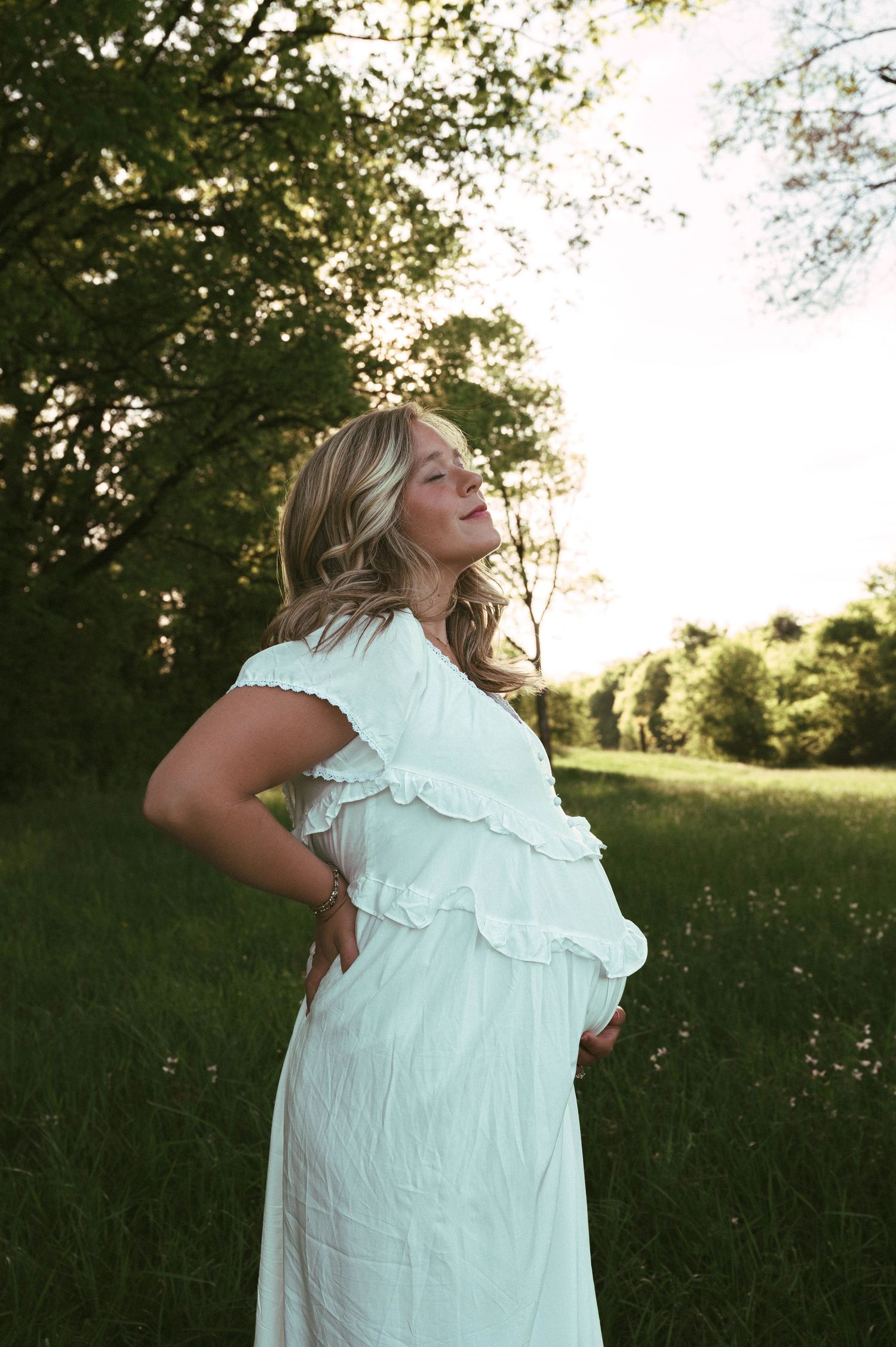 Pregnant woman in white dress standing in green field, golden hour sunlight, maternity photography outdoors.