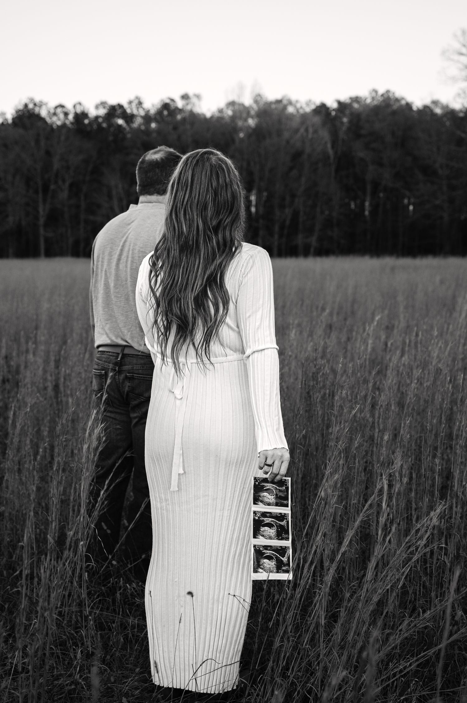 Outdoor maternity photos in tall grass field showing couple holding ultrasound image.