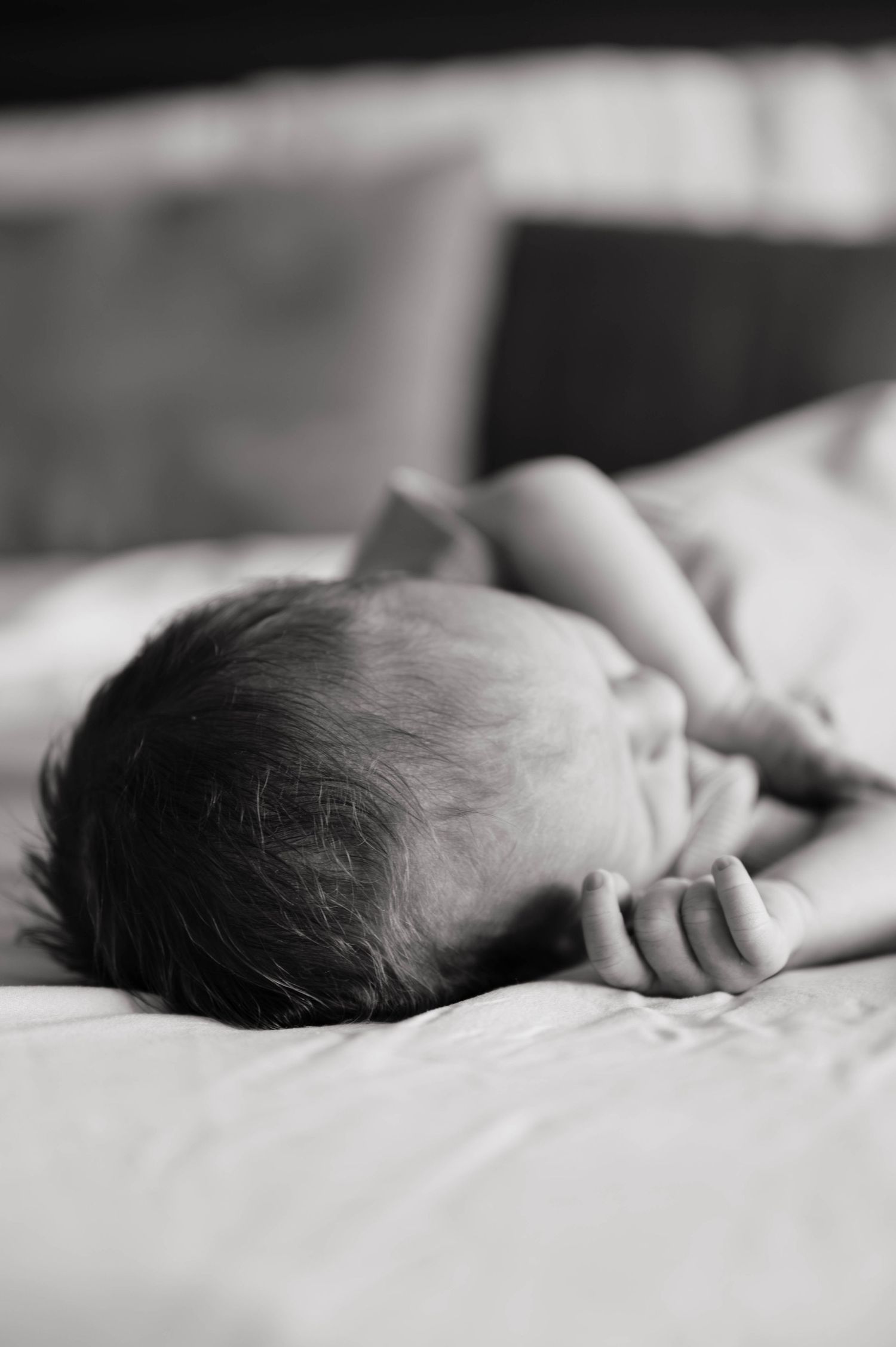 Black and white photo of a newborn baby lying on a white bed, showing tiny hands and dark hair.