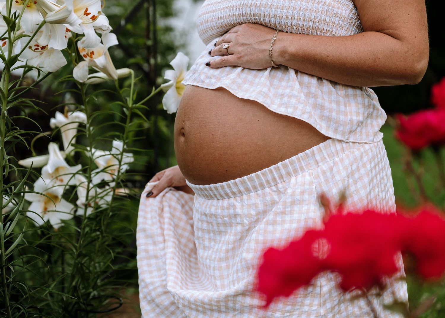 Pregnant woman in white floral dress cradling baby bump outdoors surrounded by white lilies and red roses.
