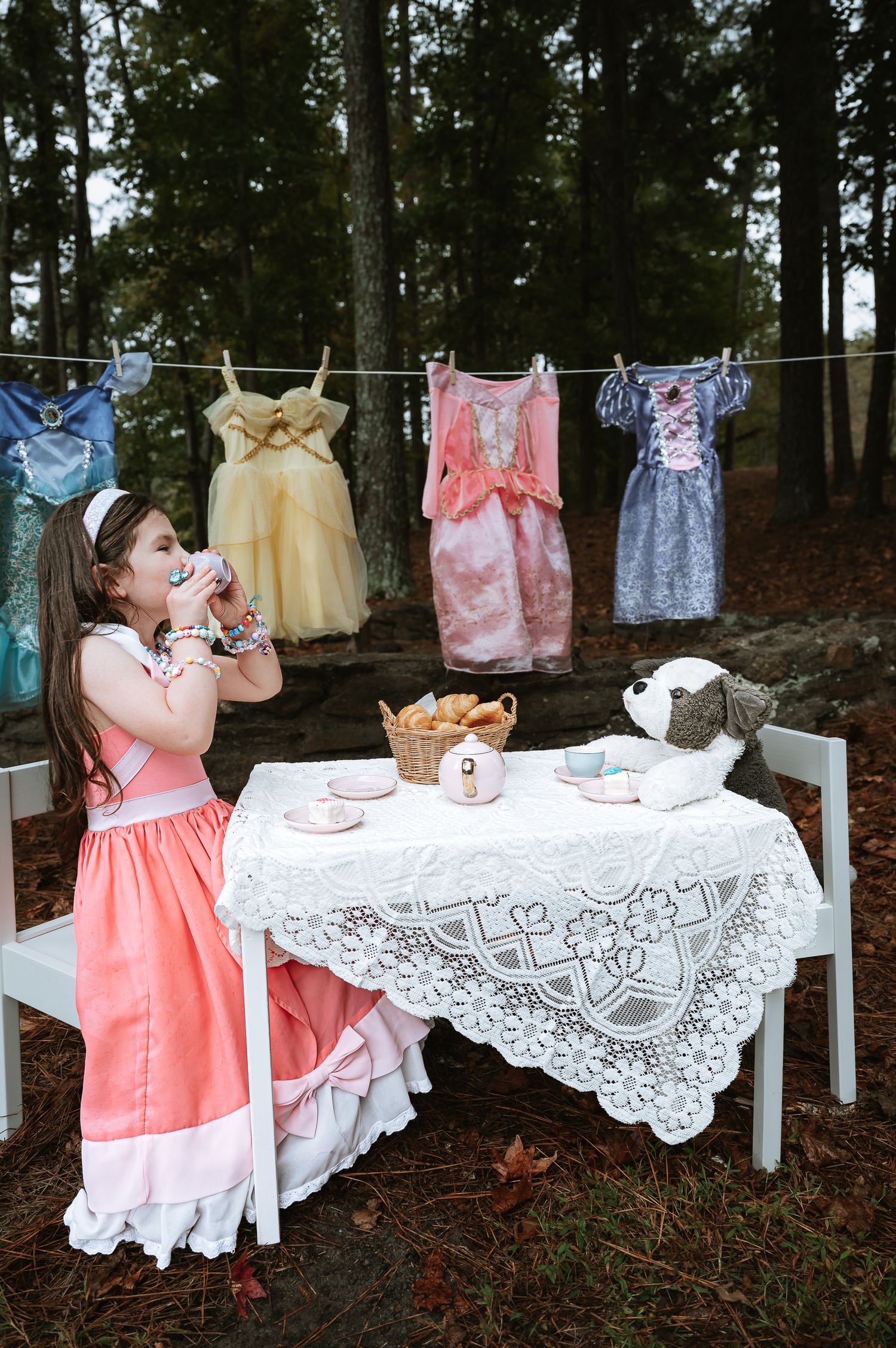 Vintage tea party setup in forest with lace tablecloth, china cups and children's dresses hanging on clothesline.