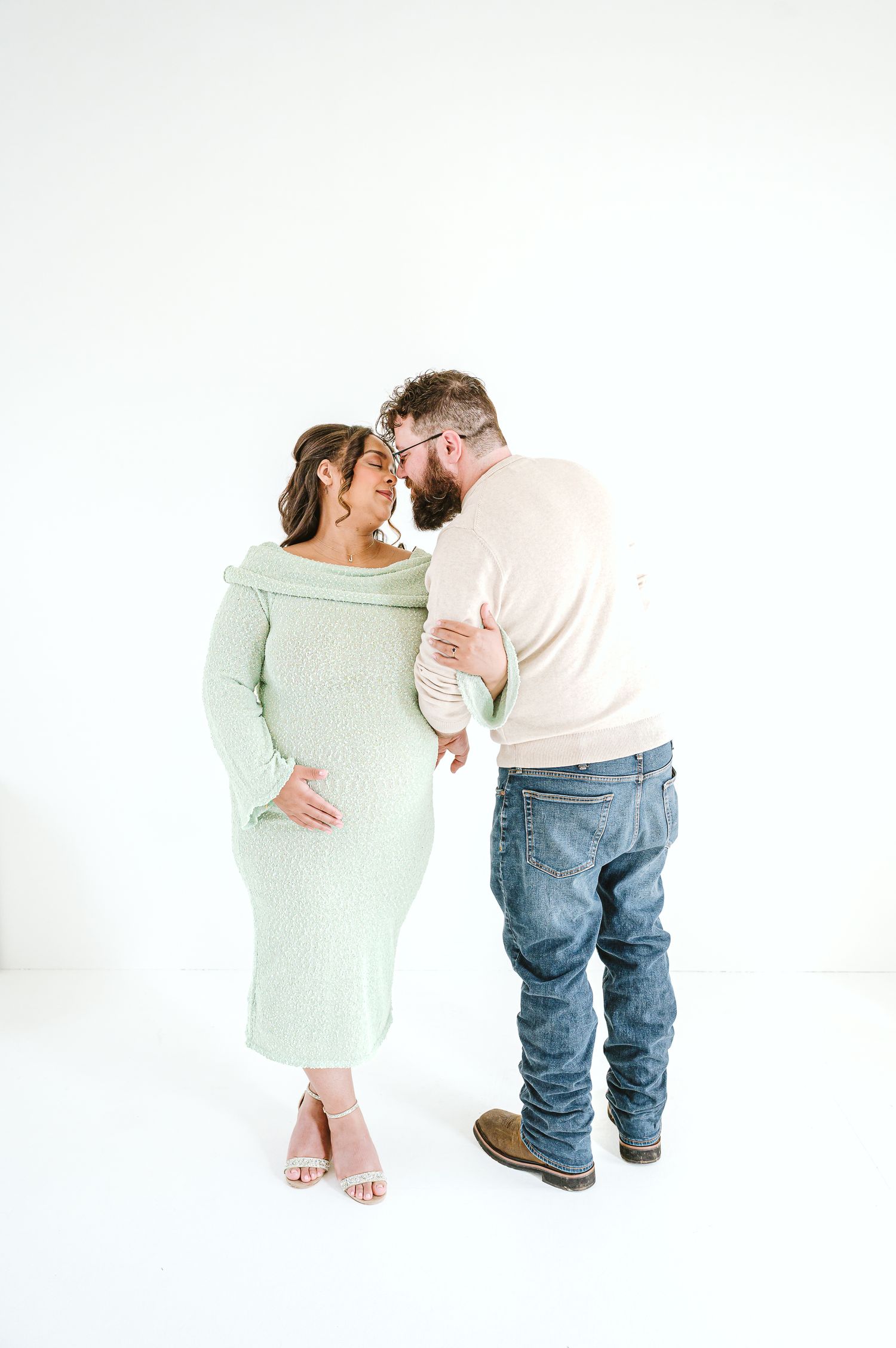 Pregnant woman in mint green dress with partner sharing a tender moment against white background.