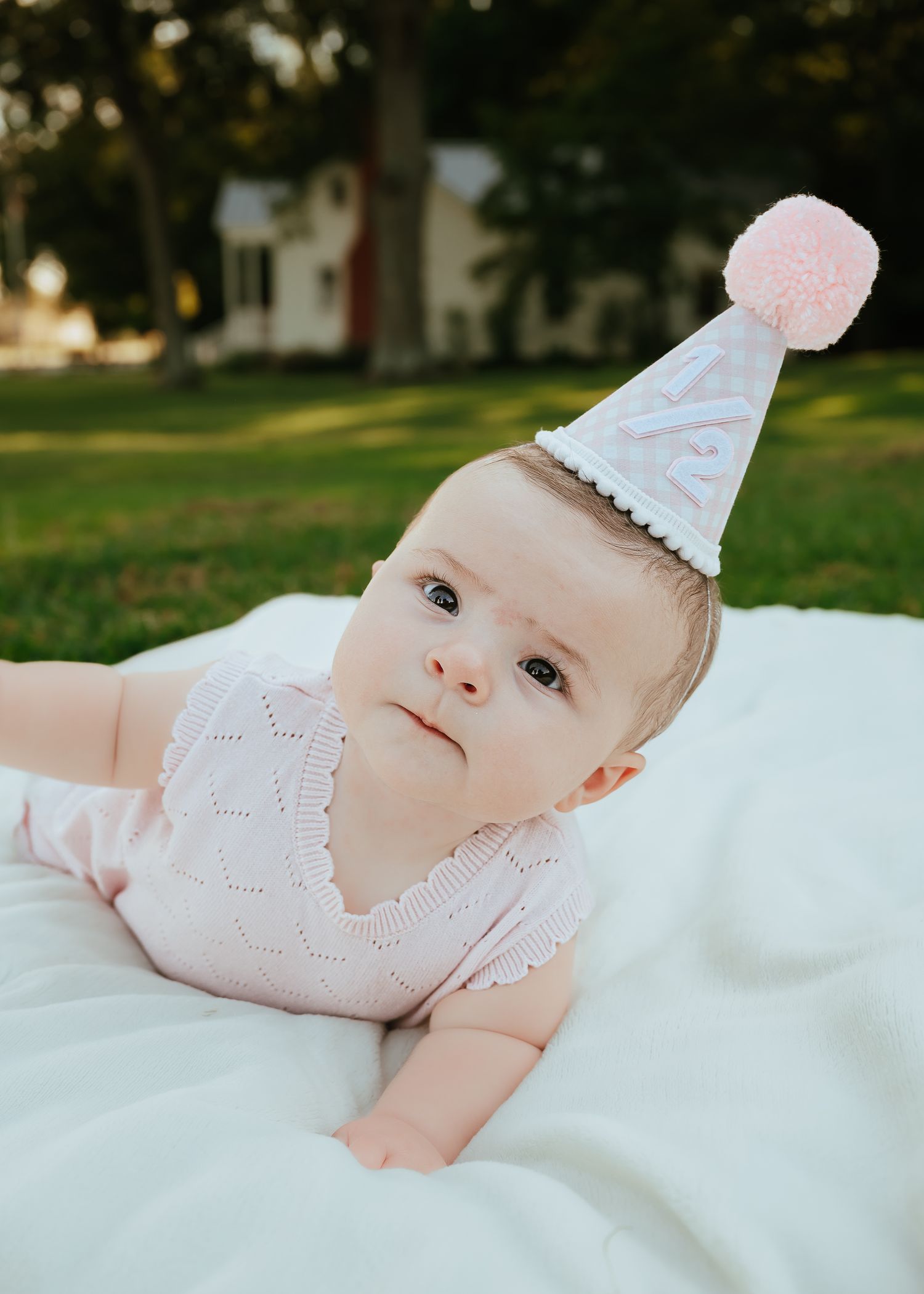 Baby wearing a pink party hat lying on a white blanket outdoors.