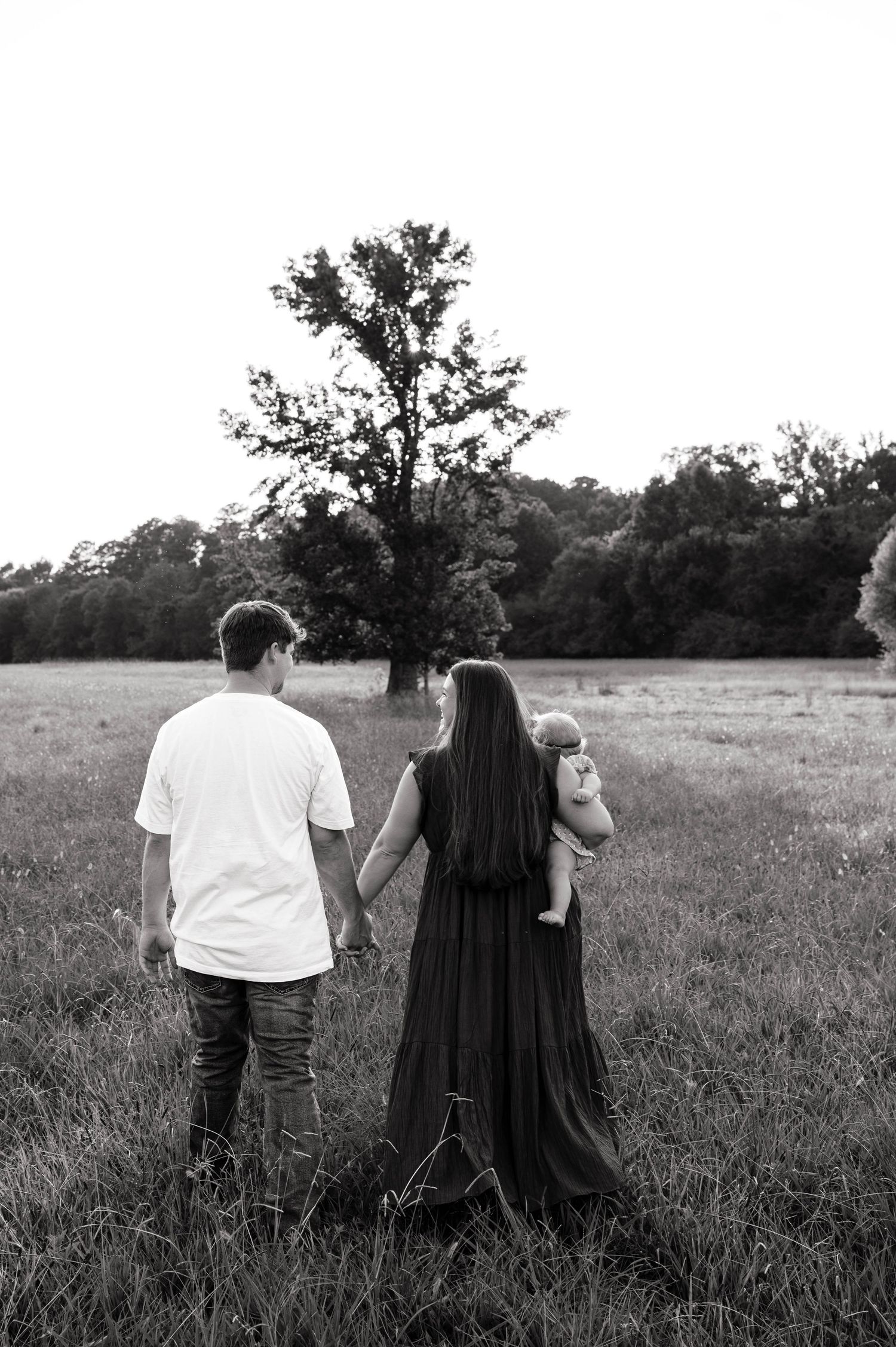 Black and white photo of a couple holding hands in a field, woman carrying a baby, with trees in the background.