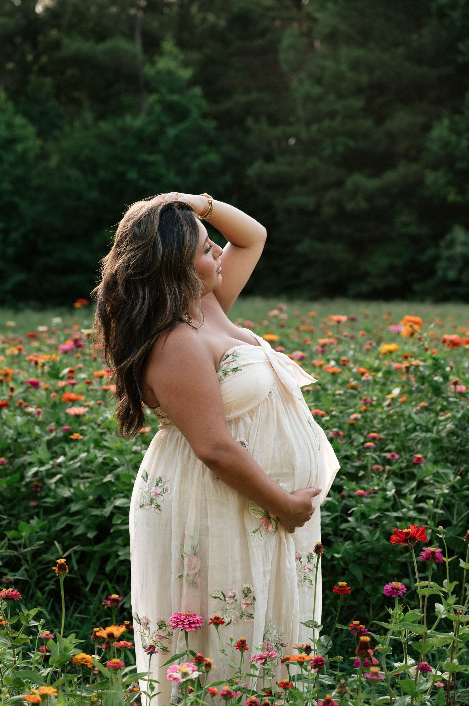 Pregnant woman in floral cream dress cradling baby bump in colorful zinnia flower field.