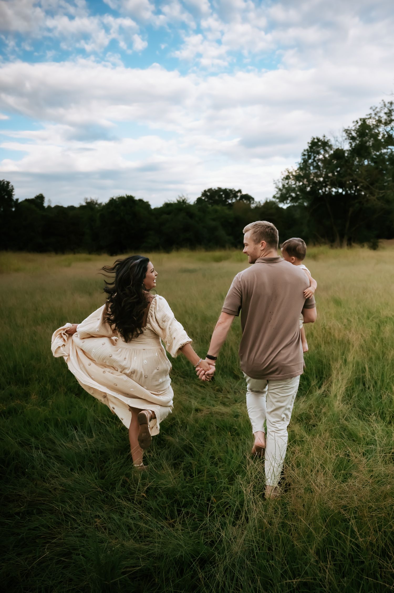 A couple walking hand in hand through a sunlit meadow with flowing dress and casual attire.