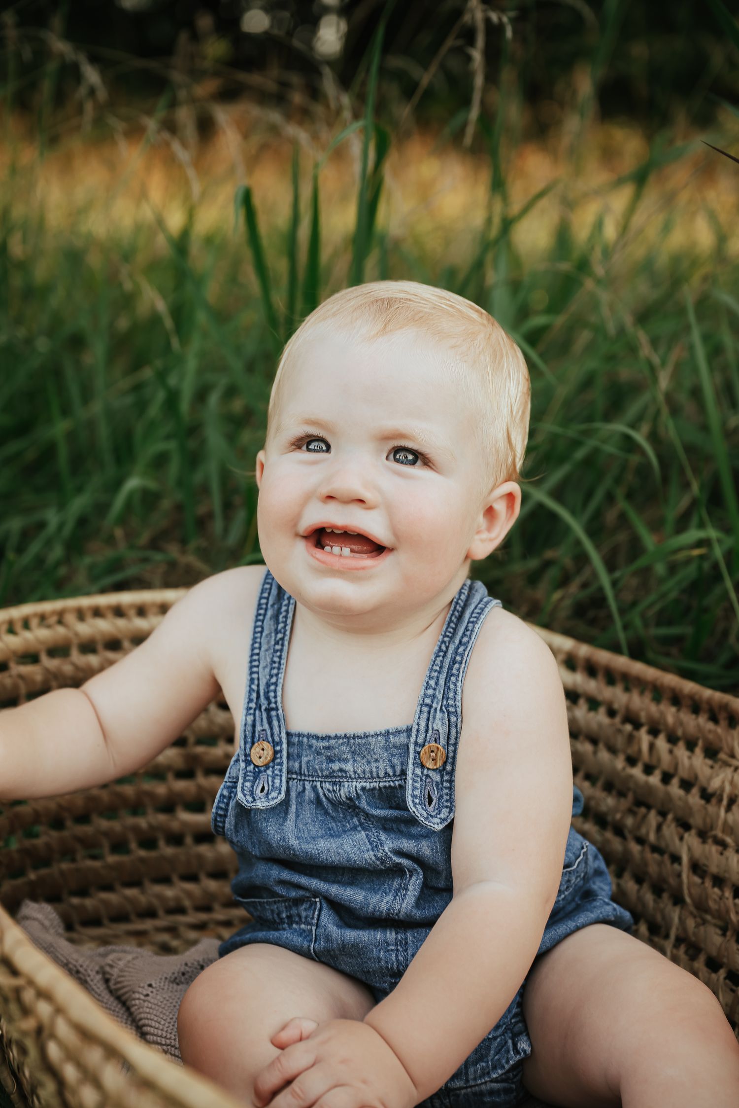 Smiling baby in denim overalls sitting in wicker basket outdoors.