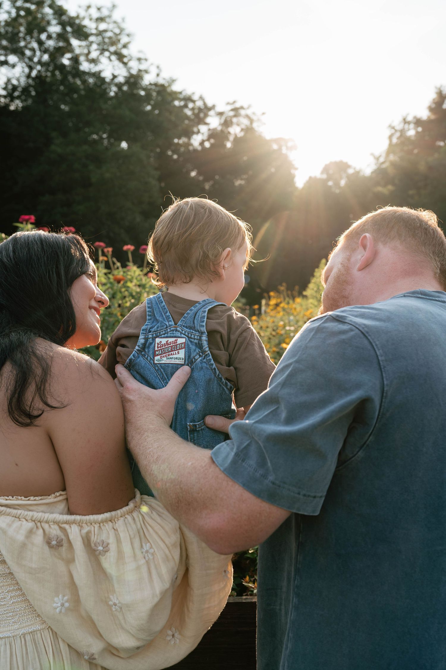 Family of three enjoying golden hour sunset in a flower garden, parents holding toddler in denim overalls.