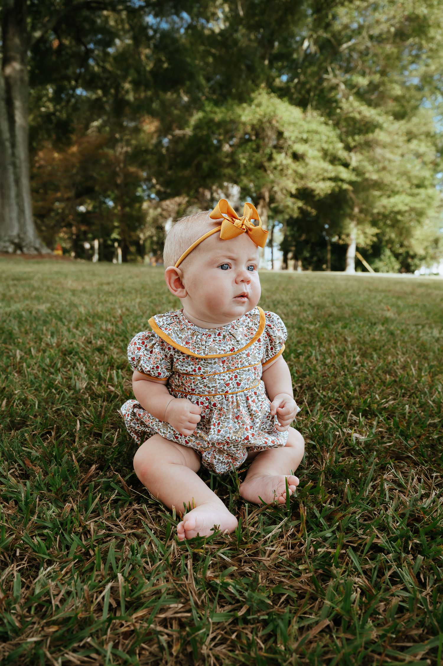 Baby wearing floral dress sitting on grass in outdoor autumn setting.