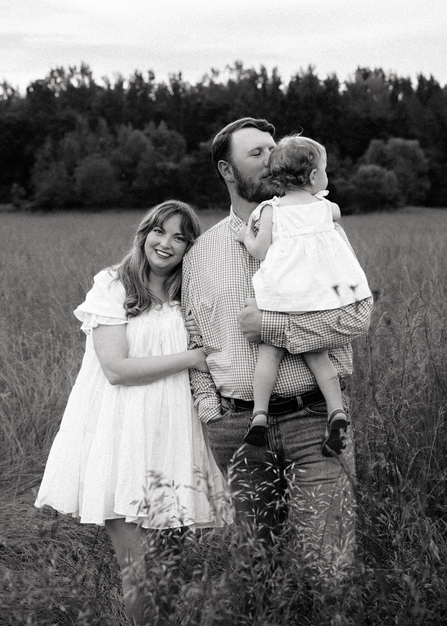 Black and white family photo of a couple and toddler girl in a field, father kissing child while mother smiles.
