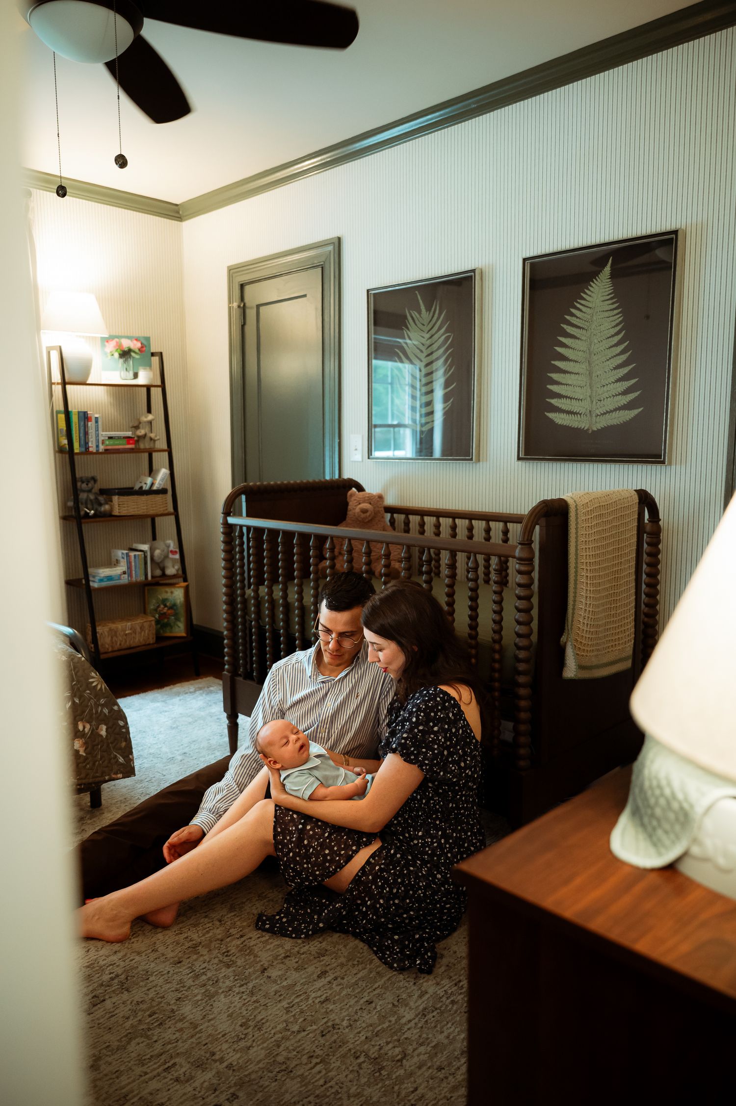 A nursery room with fern artwork and a dark wooden crib where two people sit on the floor.