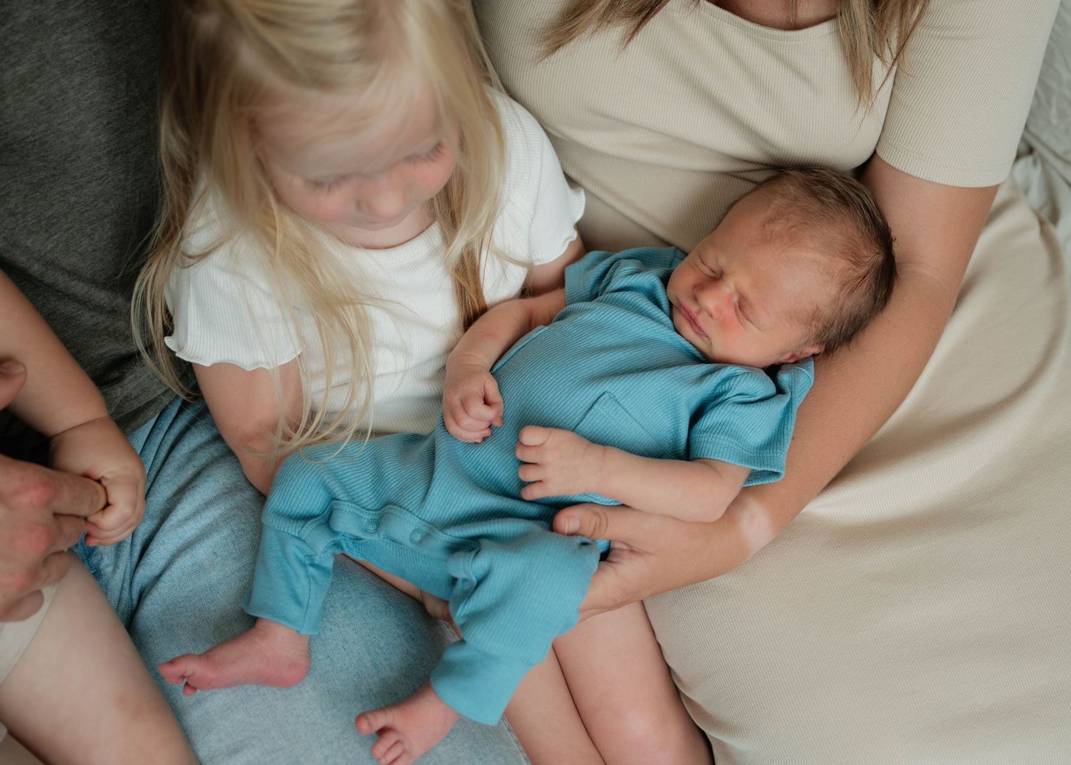 A toddler with blonde hair sits beside a sleeping newborn in a teal outfit, held by a woman in a beige top.