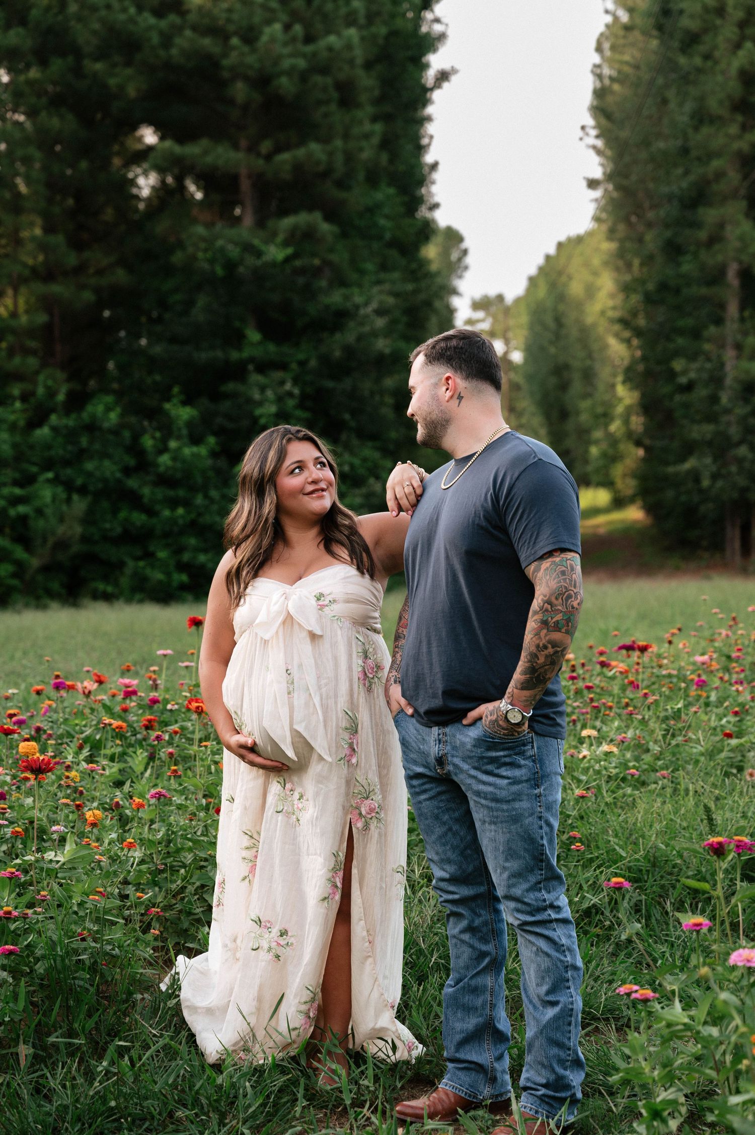 Pregnant woman in floral maternity gown stands with tattooed partner in colorful flower field surrounded by tall trees.