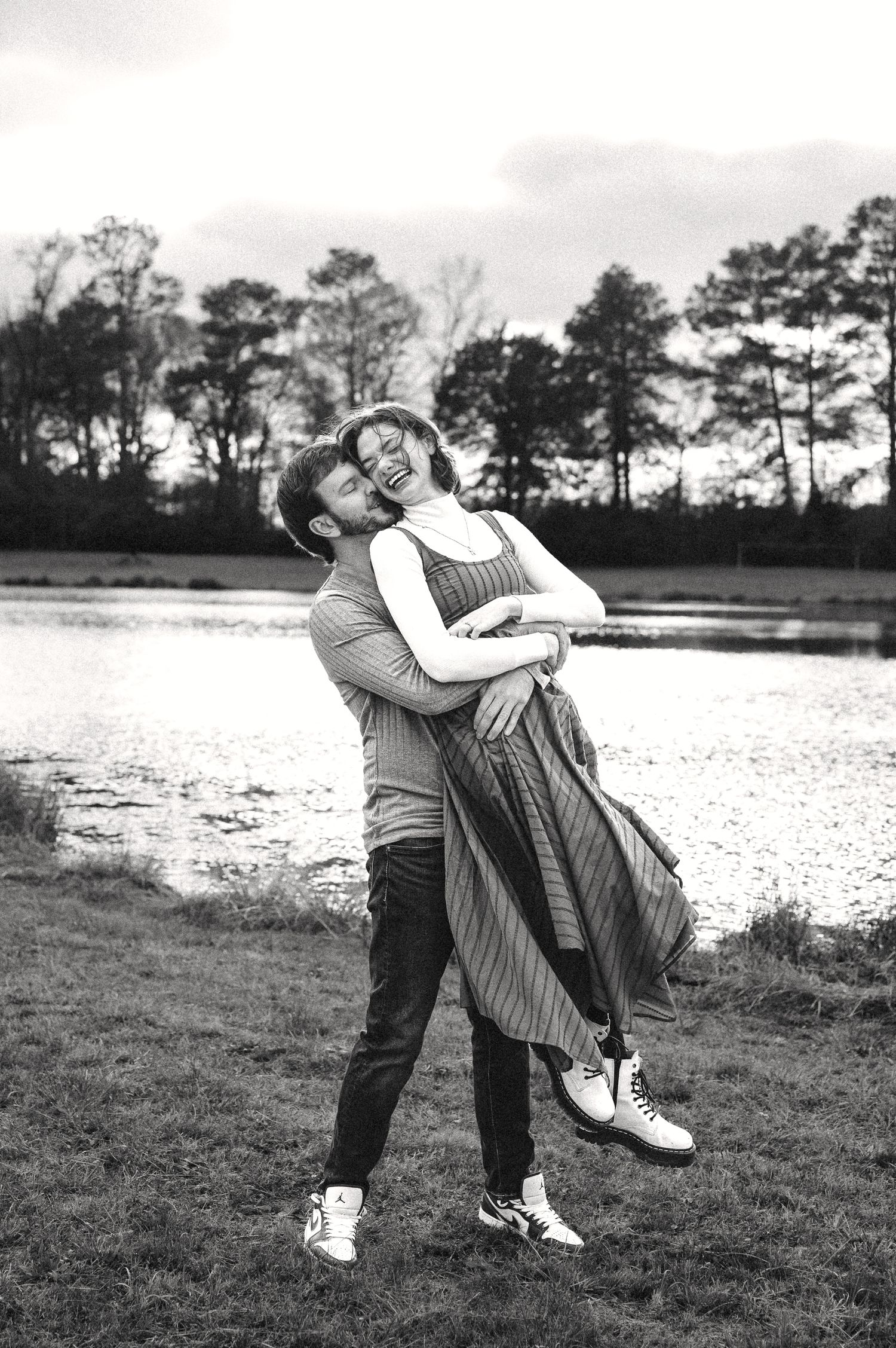 A black and white photo of a couple in a playful lift pose by a lake with trees in the background.