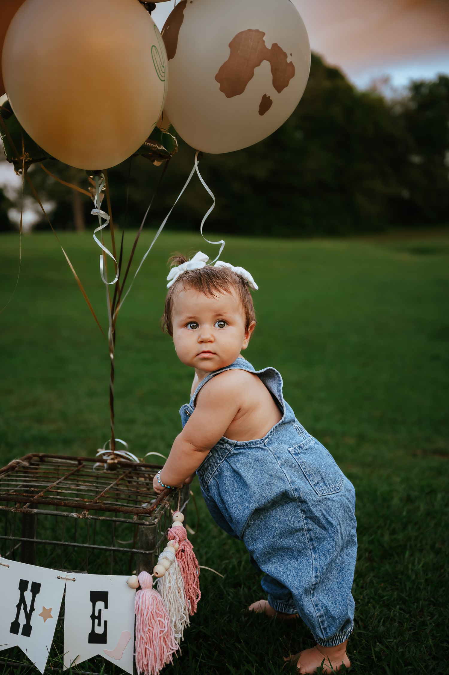 Baby in denim overalls poses with balloons outdoors during a photo session.